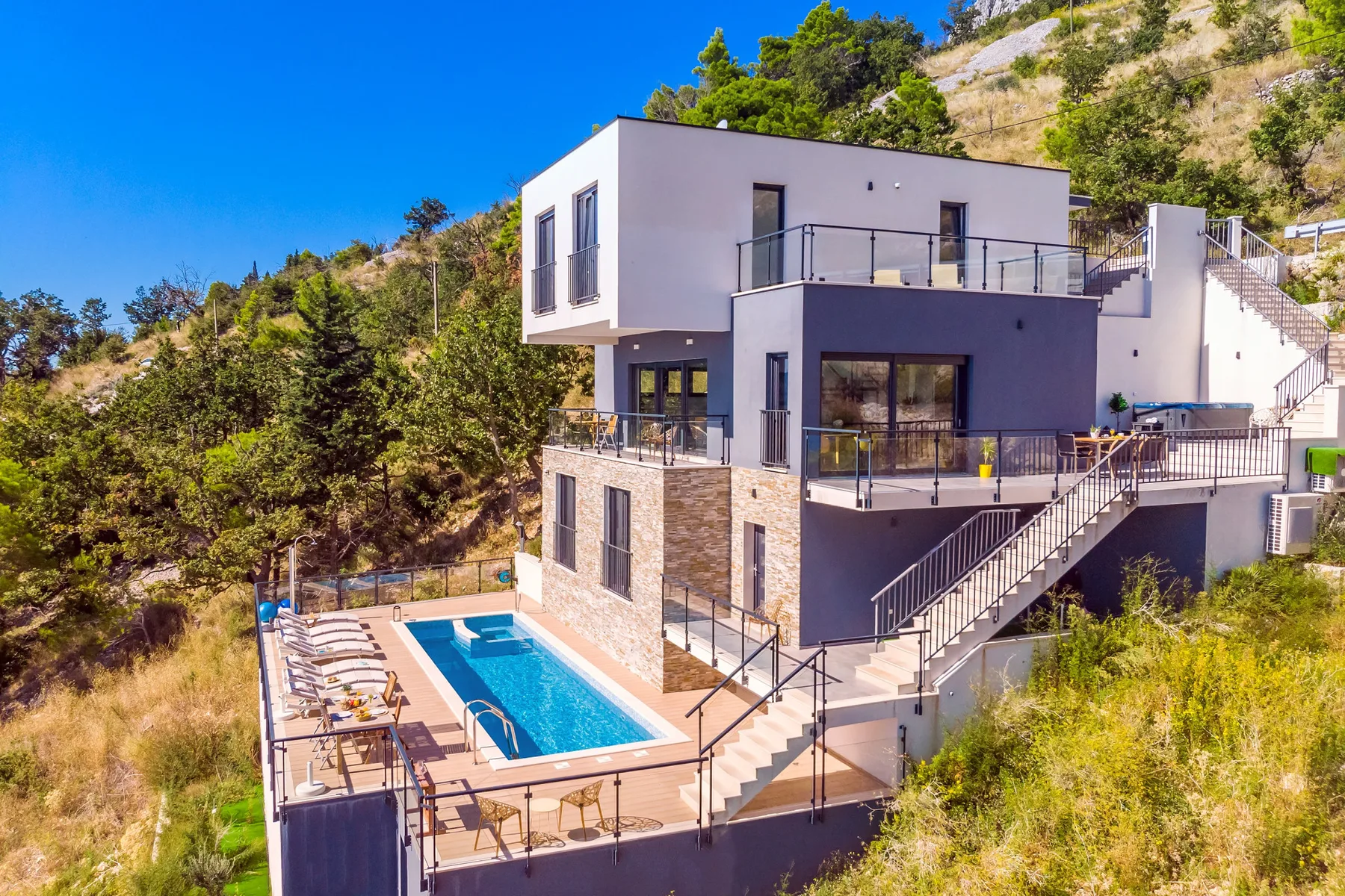 A modern hillside house overlooks an outdoor swimming pool and surrounding greenery.