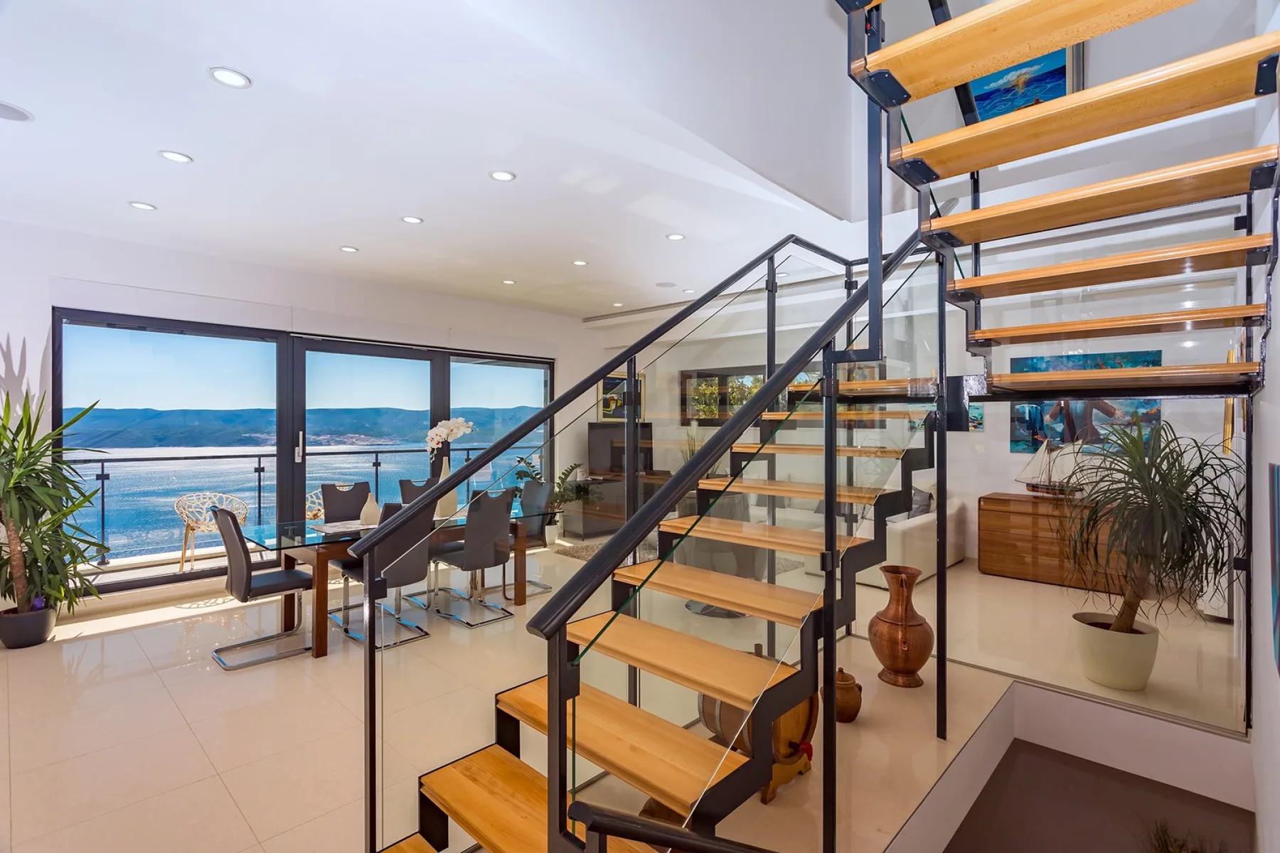 A modern living and dining area features a staircase and large windows overlooking the water.