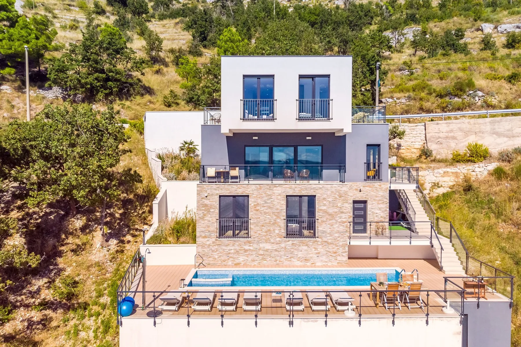 A modern hillside house overlooks a terrace with a swimming pool.