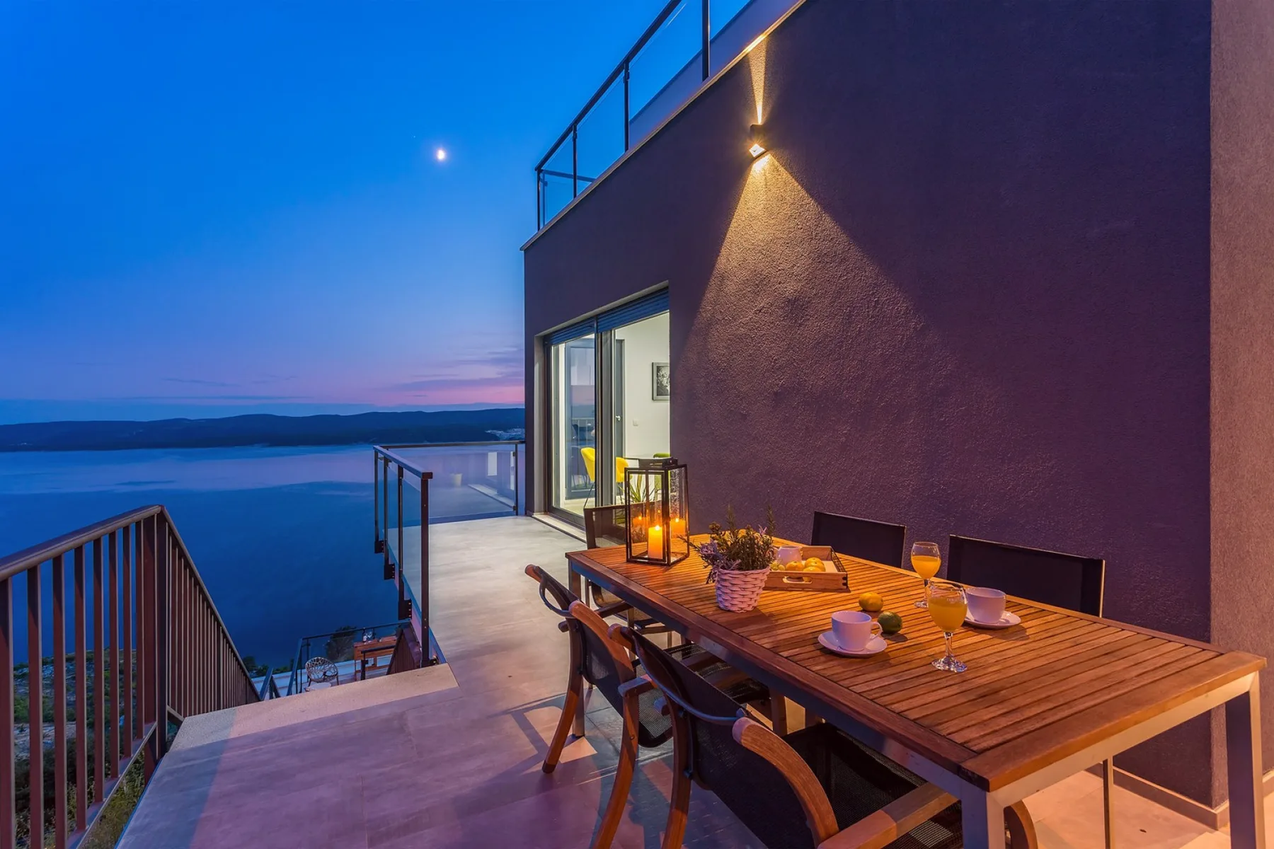 An outdoor terrace with a dining table overlooks the water at dusk.