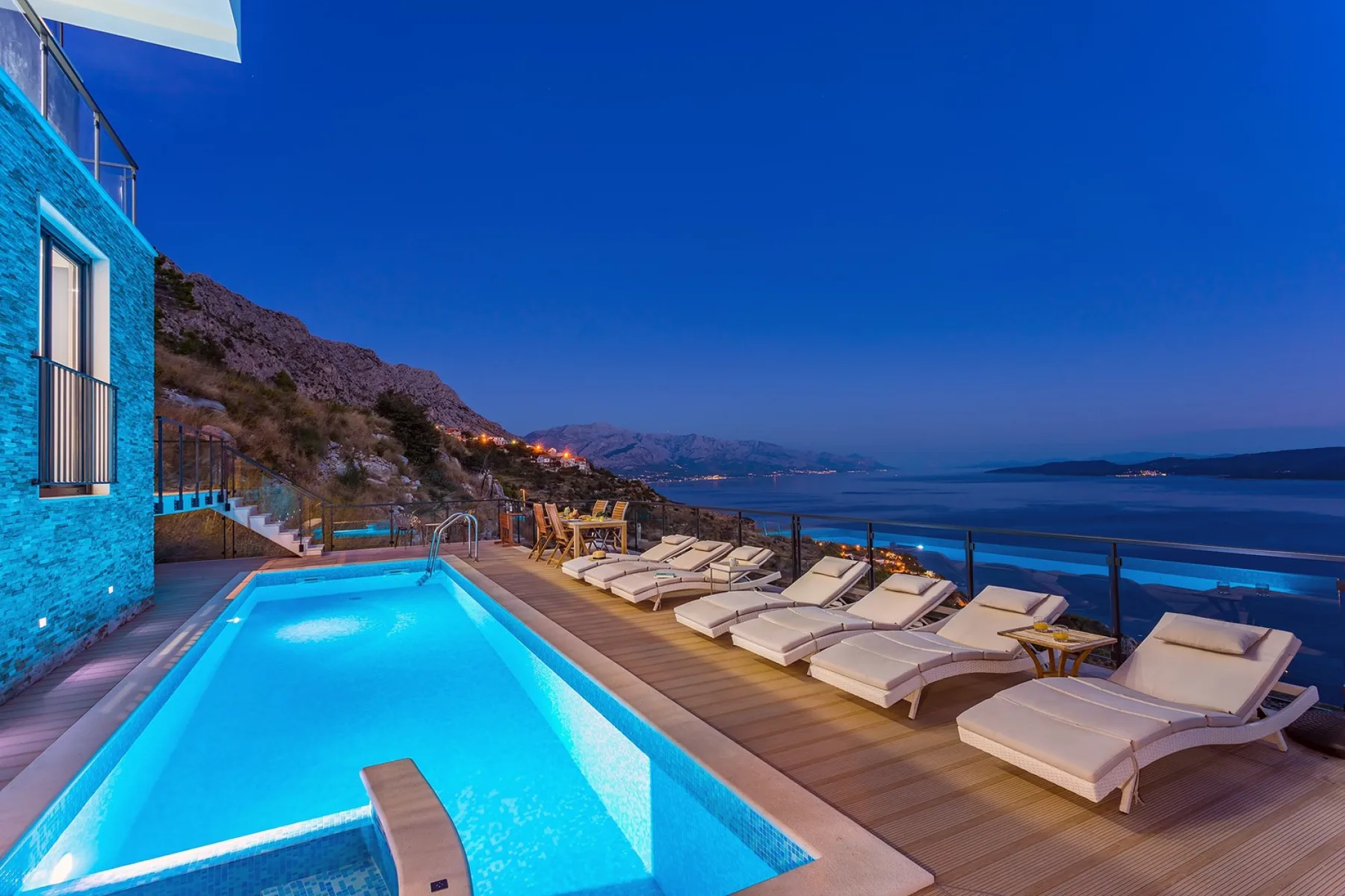 A lit swimming pool and lounge seating overlook the sea at dusk.
