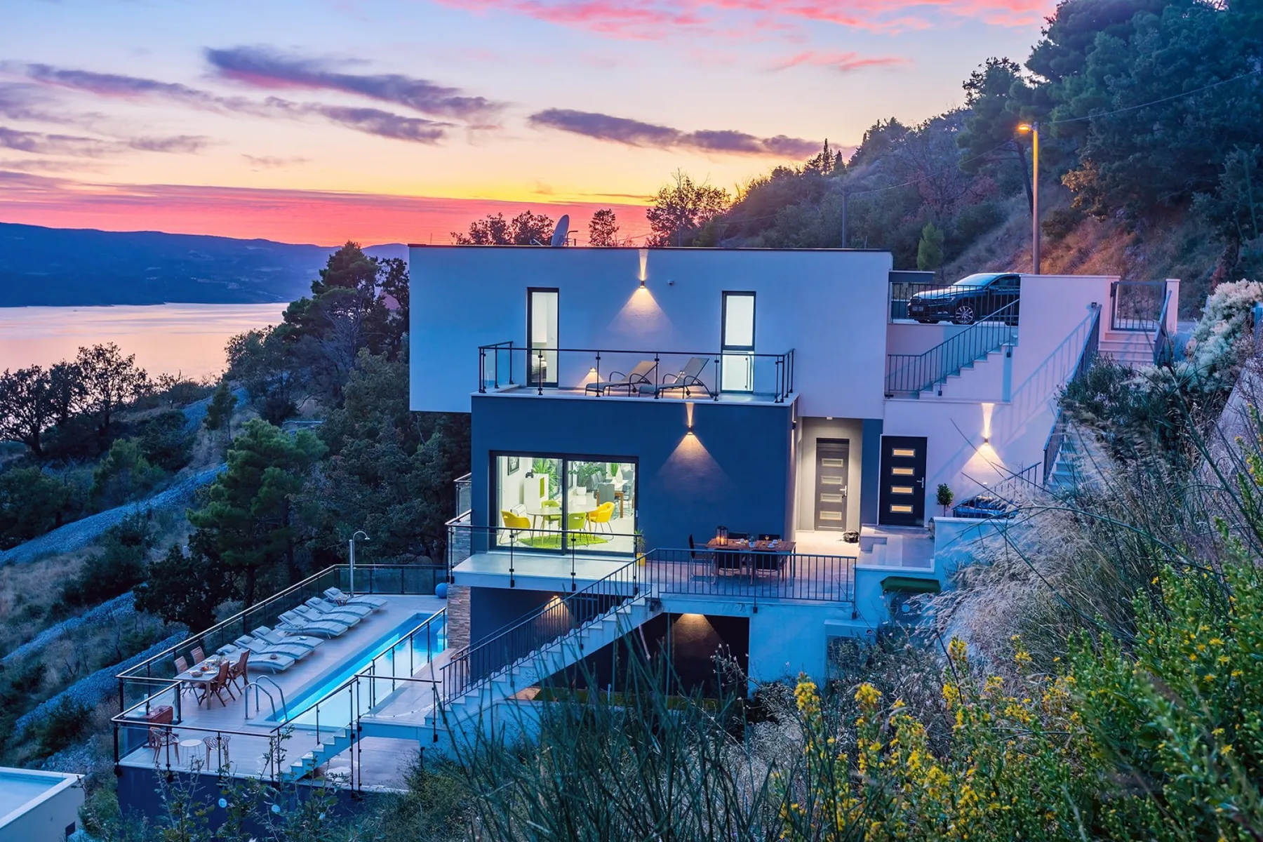 A modern hillside house overlooks a lake beneath a colorful sunset sky.