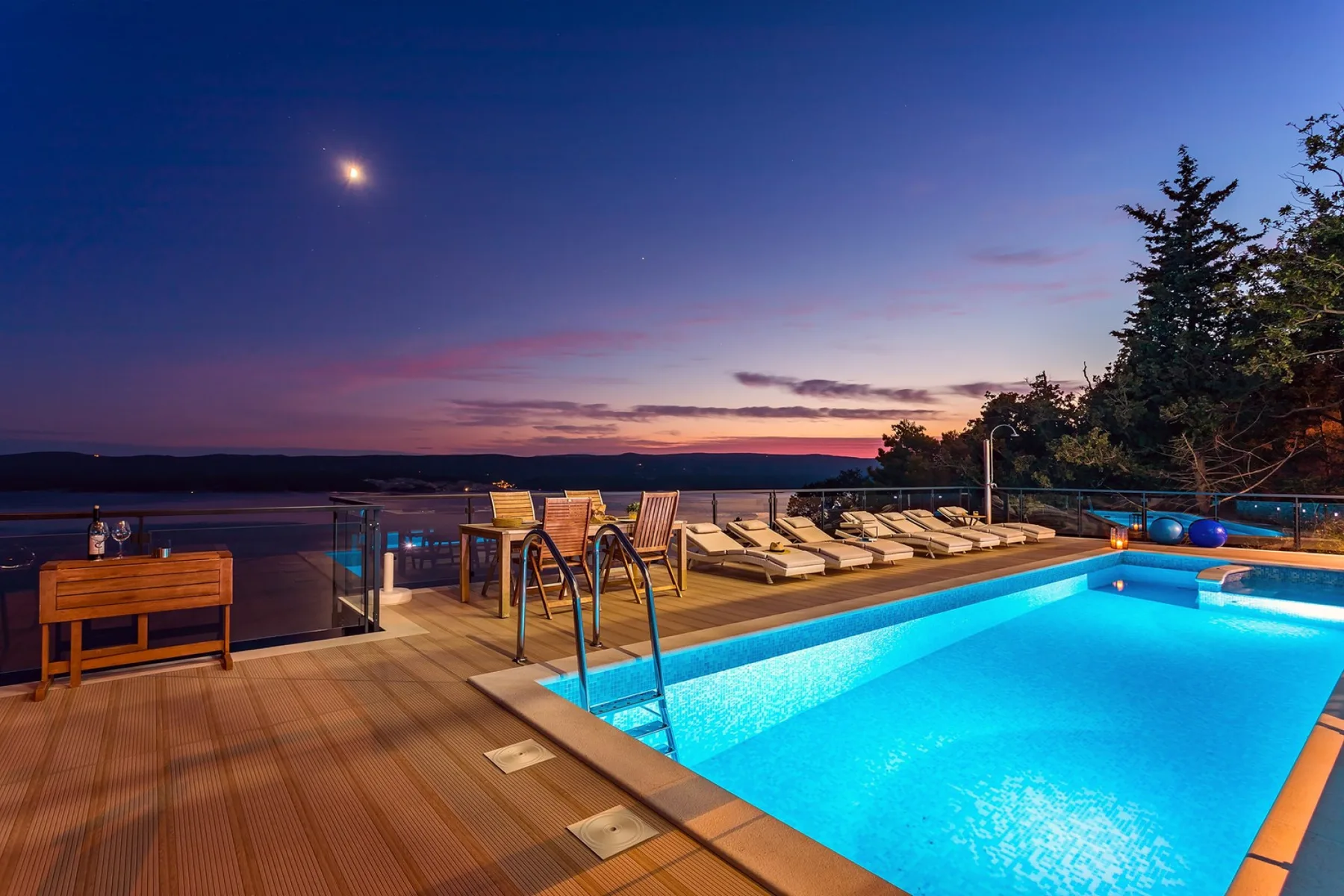 An outdoor pool and deck overlook a lake beneath the colorful evening sky.