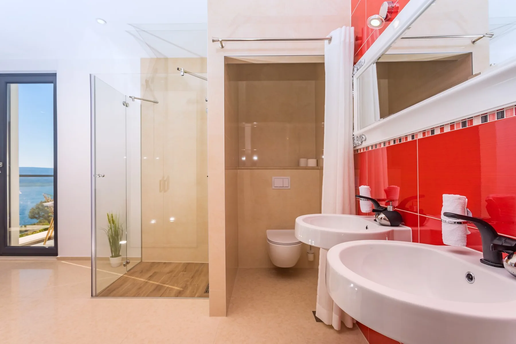 A bright bathroom with a glass shower, wall-mounted toilet, and sinks beside a red tiled wall.