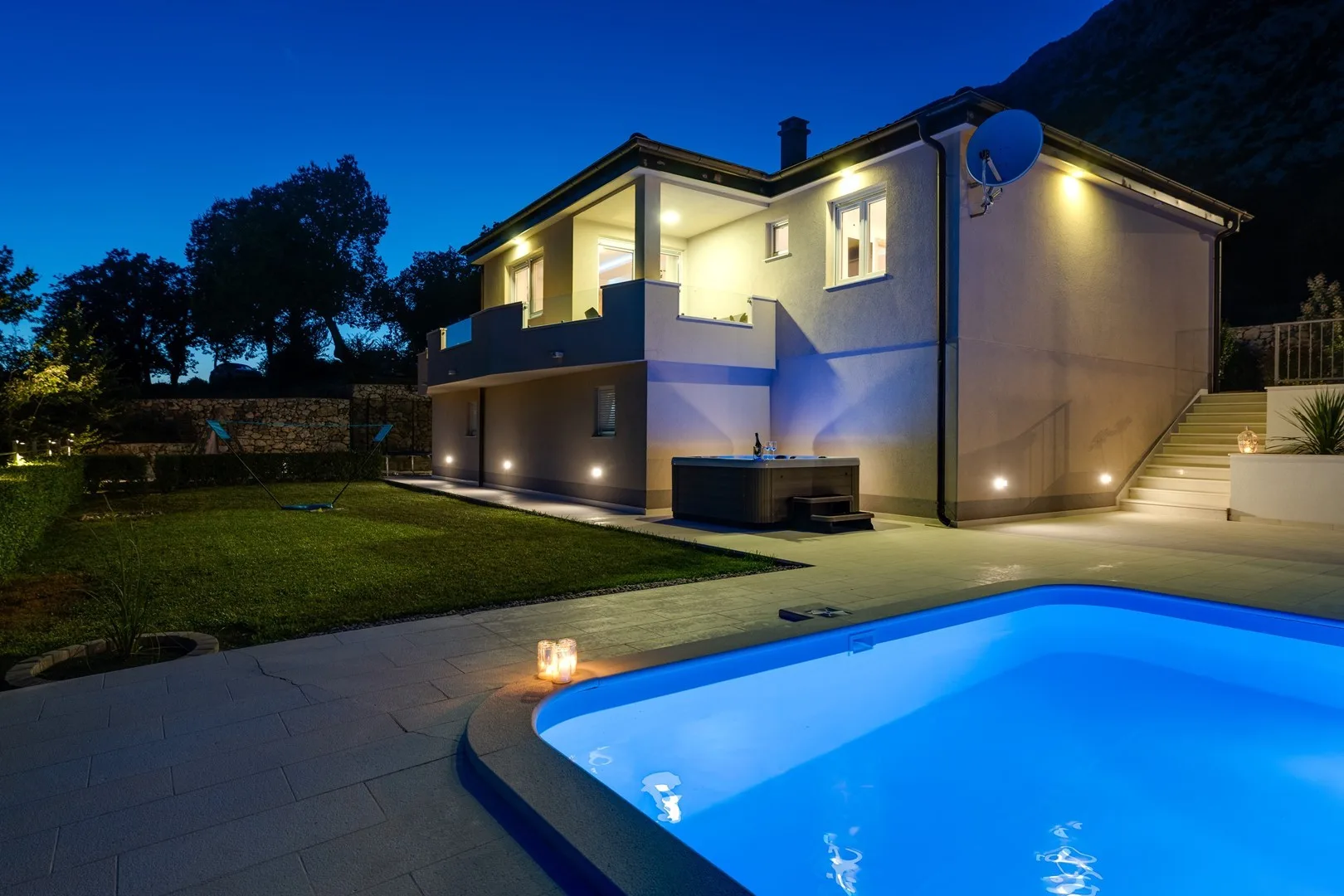 A modern house and illuminated swimming pool stand in a landscaped yard at dusk.