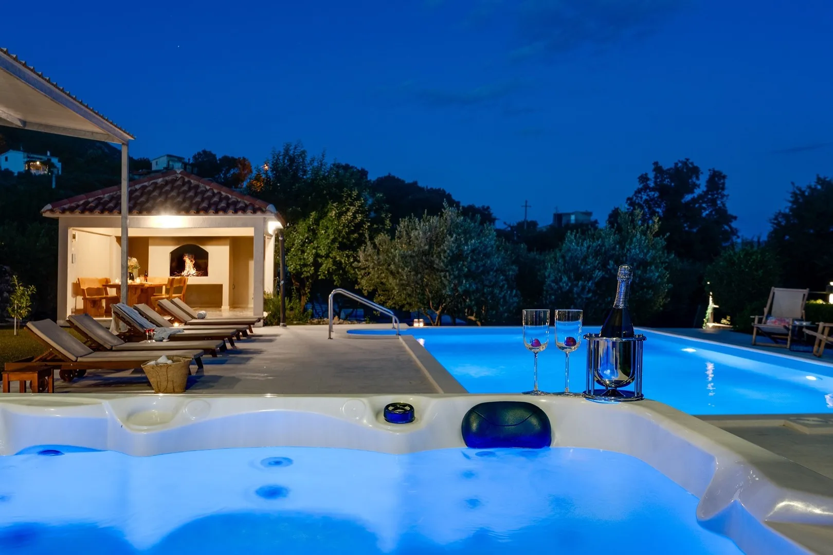 An outdoor pool and illuminated hot tub sit beside a covered patio at dusk.
