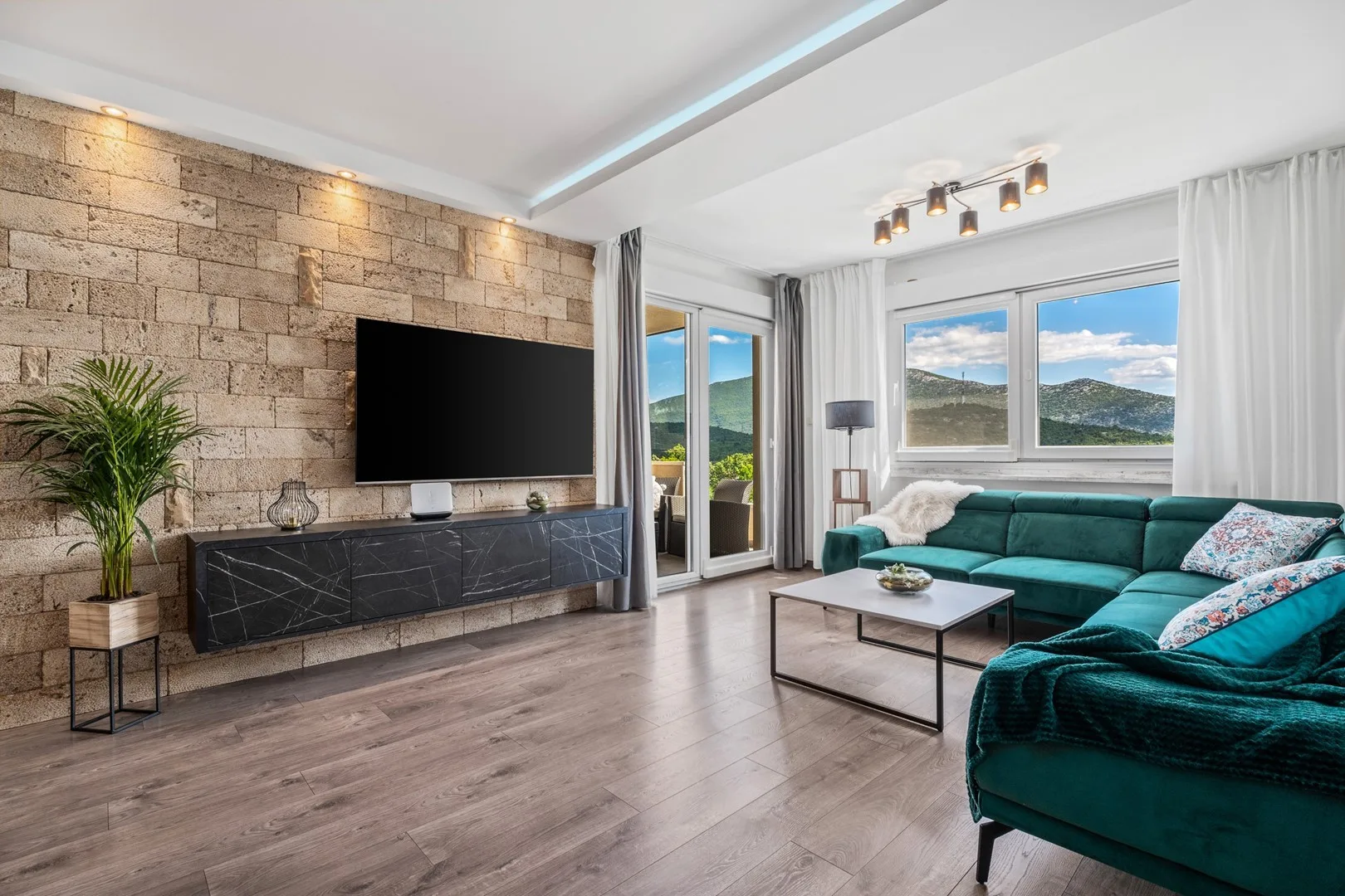 A bright living room with teal seating, a television, and large windows overlooking hills.