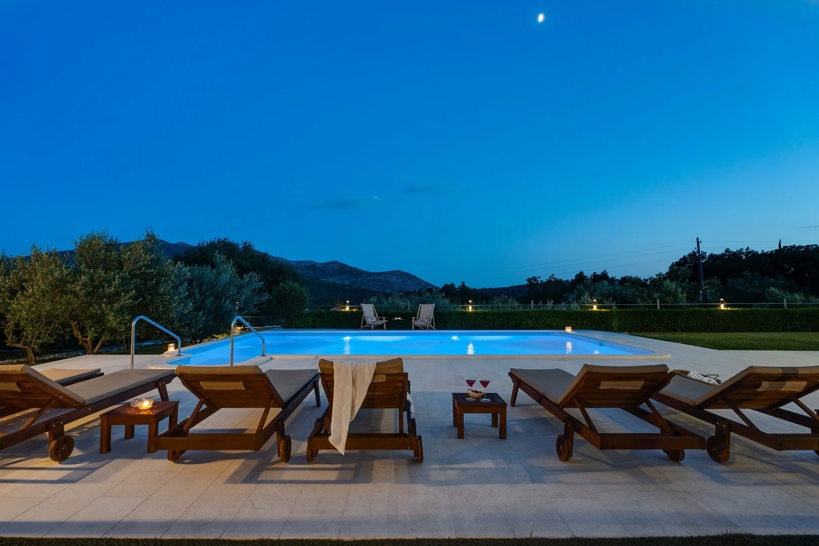 A swimming pool at dusk is surrounded by wooden loungers on a tiled patio.
