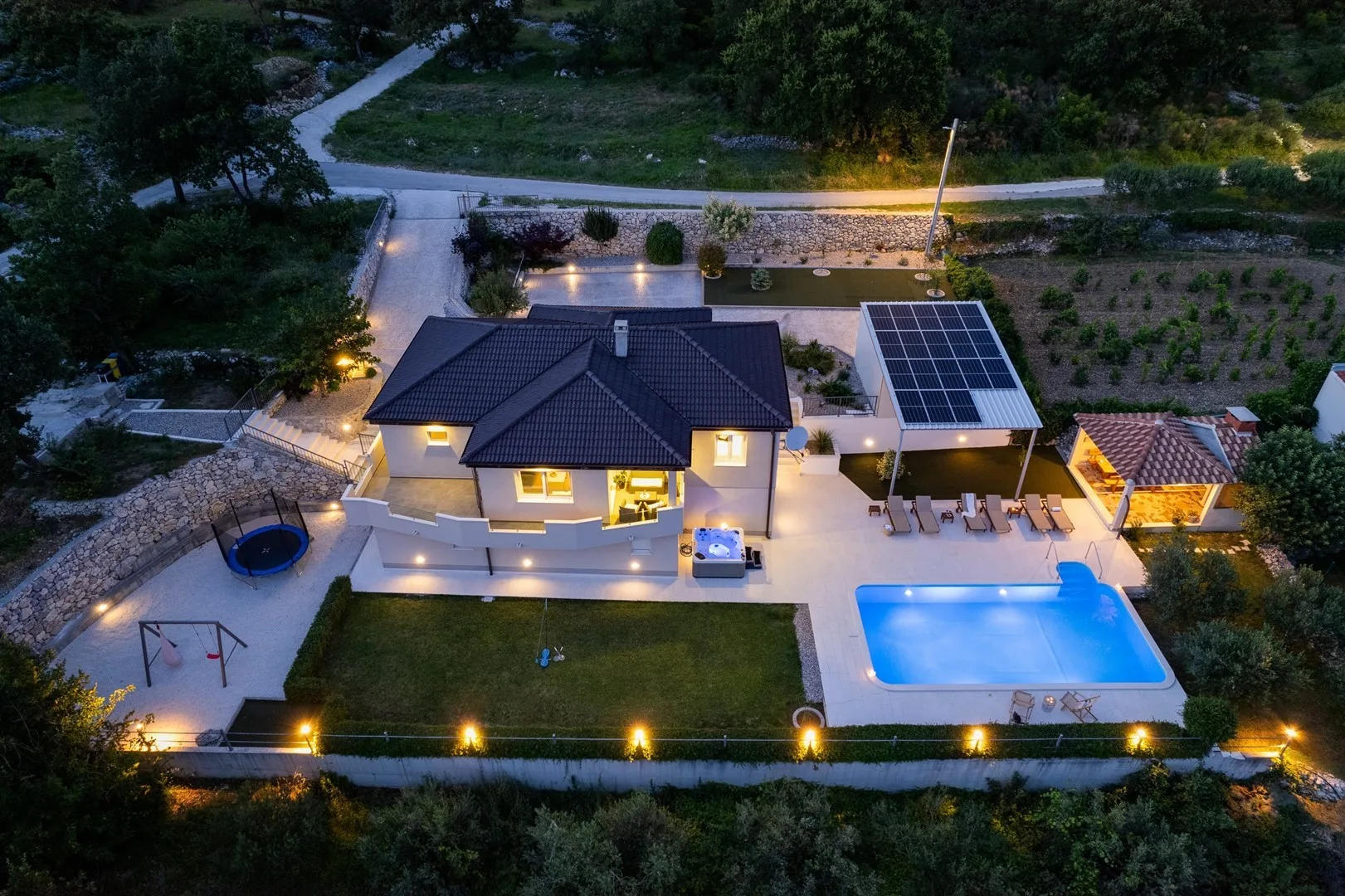 An illuminated house and swimming pool sit within a landscaped outdoor area.