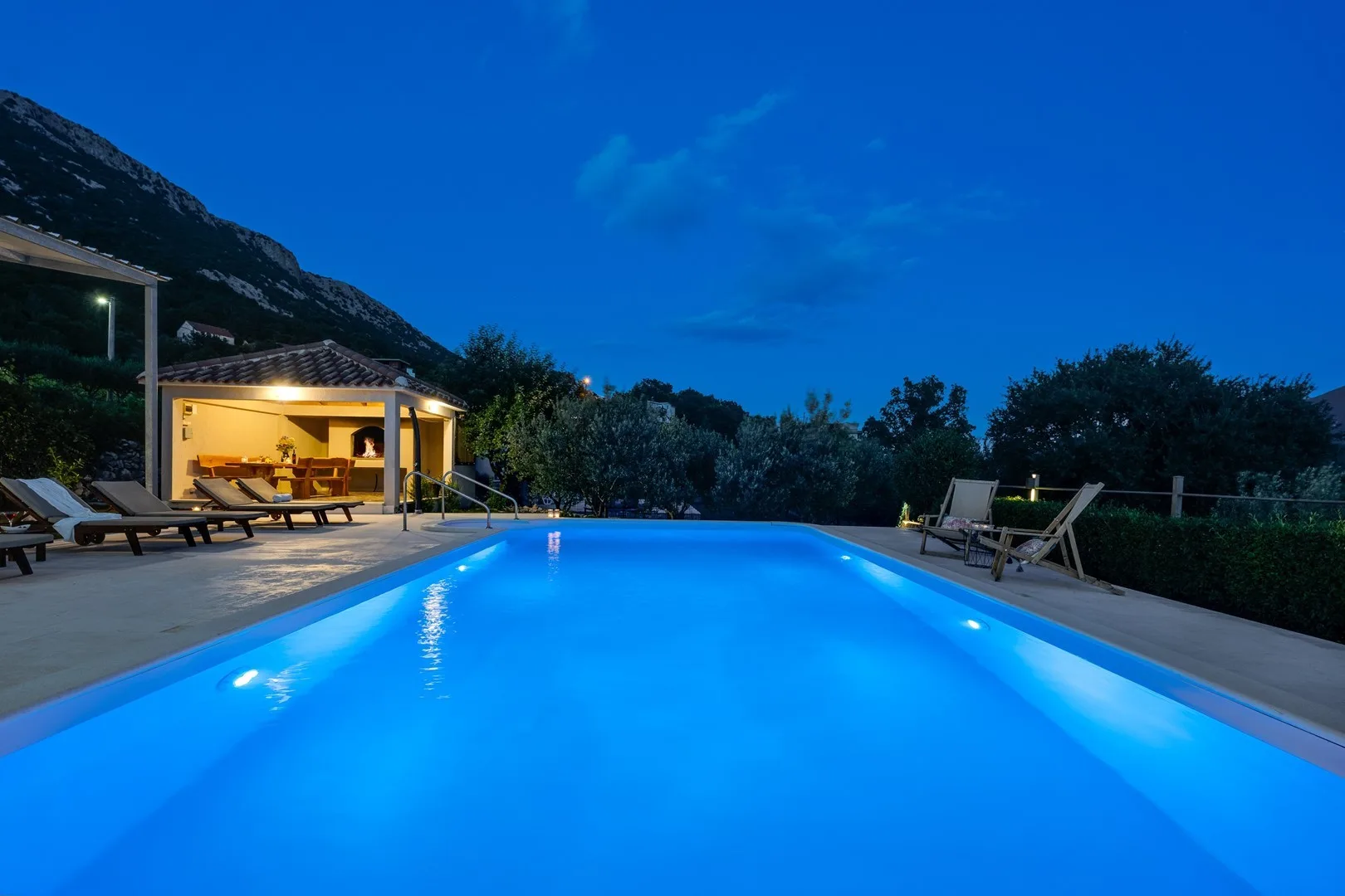 An illuminated swimming pool at dusk, with lounge seating beside it.