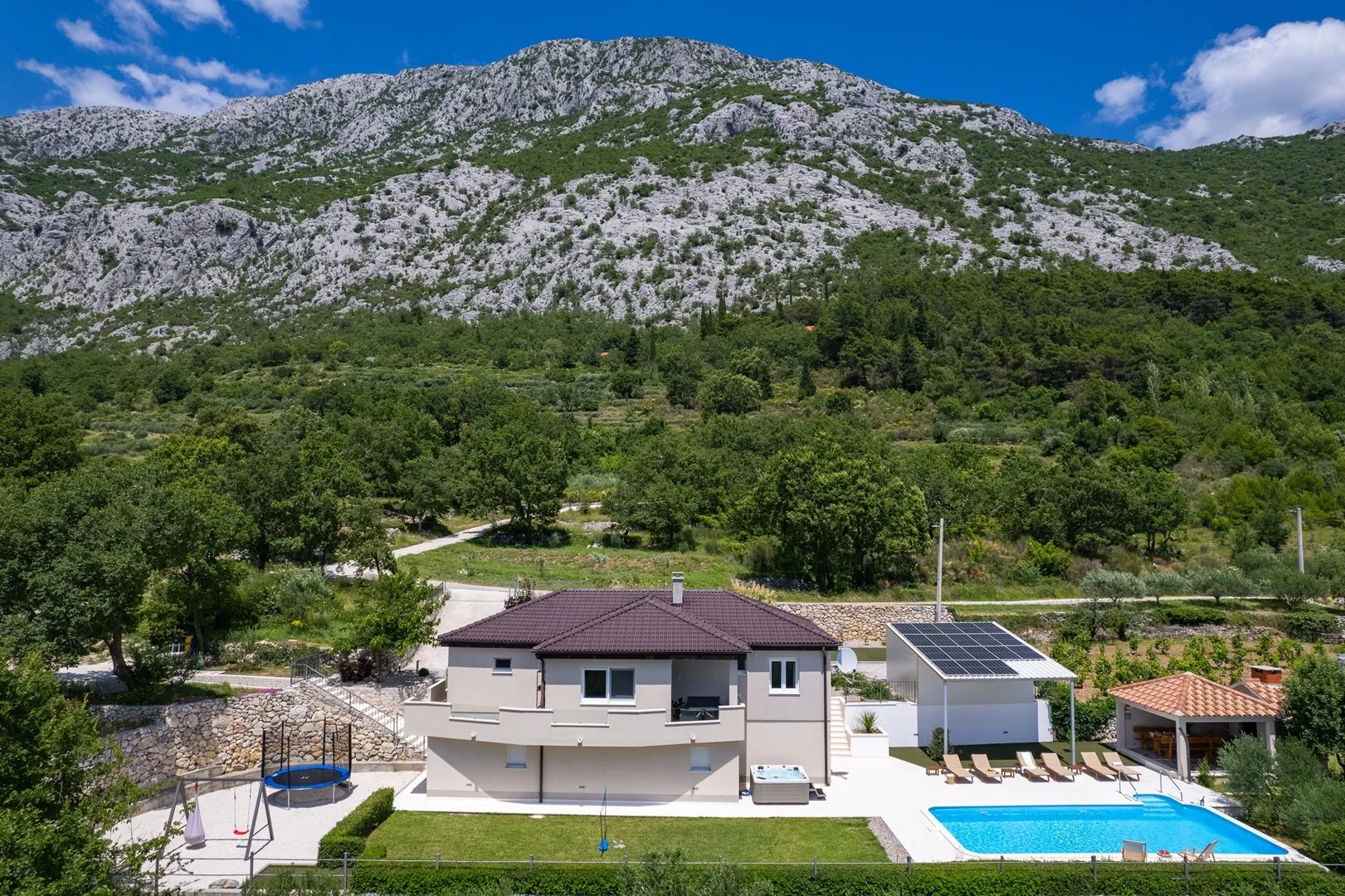 A house and swimming pool sit among trees below a rocky mountain.