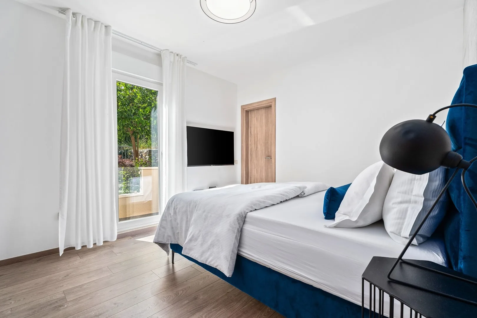 A bright bedroom with a bed, white curtains, and a large window.