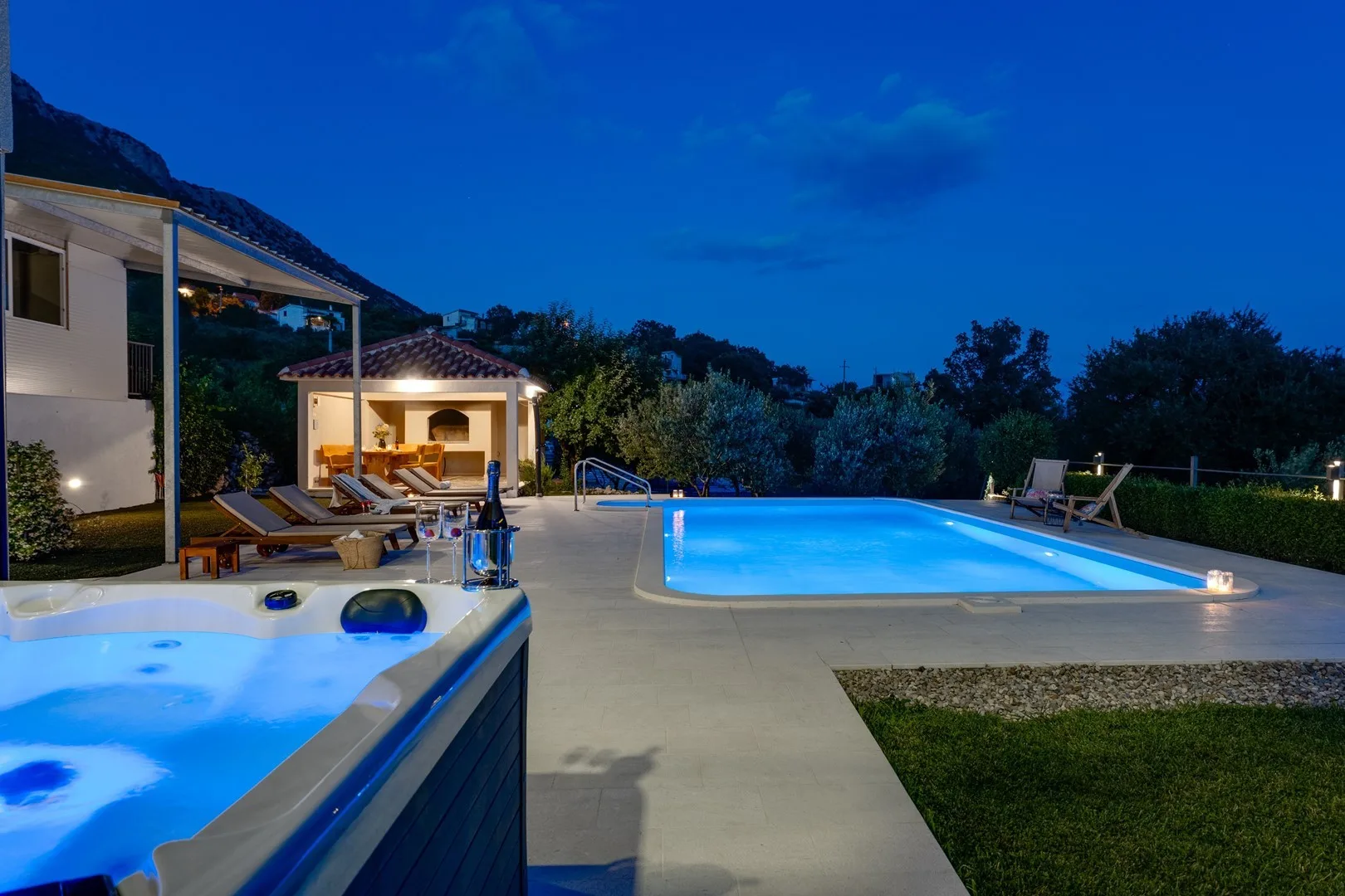 An outdoor pool and hot tub glow beside a patio at dusk.