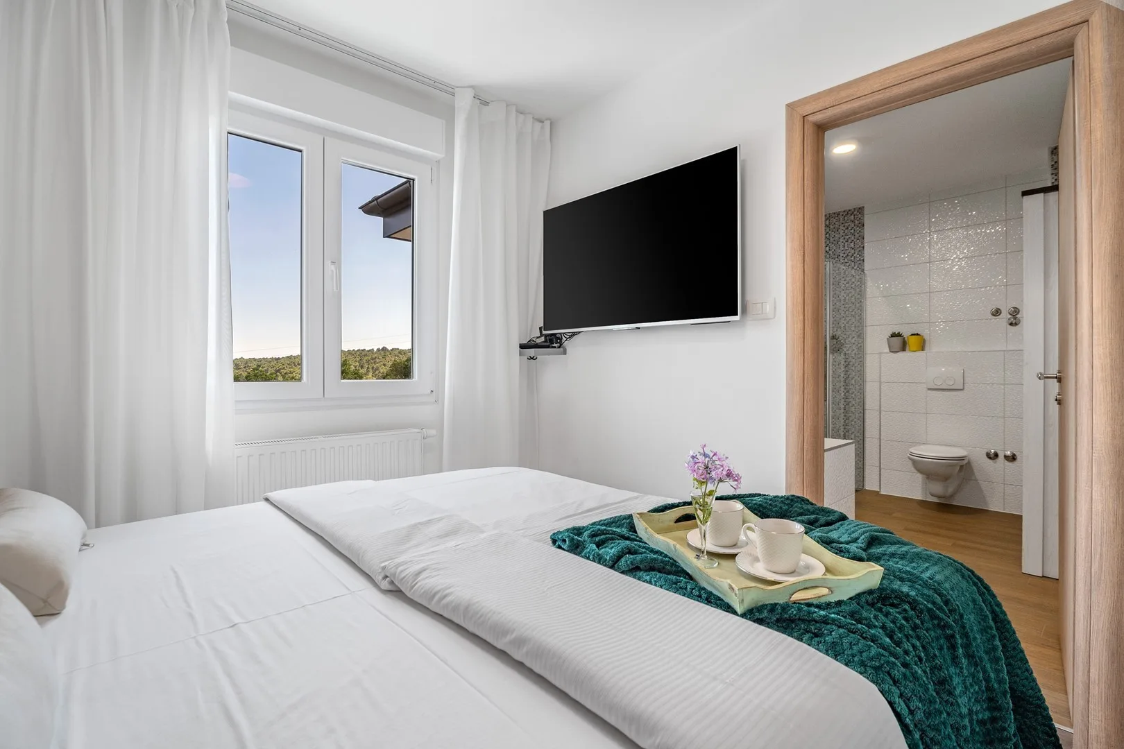 A bedroom with a white bed, a wall-mounted television, and an open bathroom doorway.