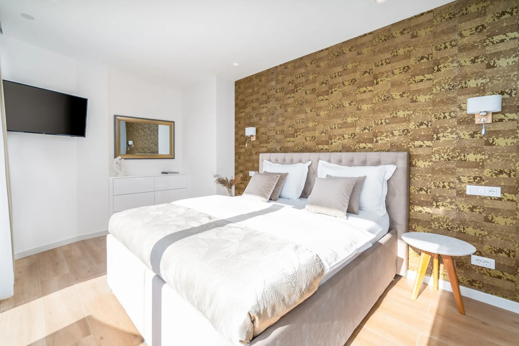A bright bedroom with a double bed, white bedding, a patterned wood accent wall, and a small bedside table.