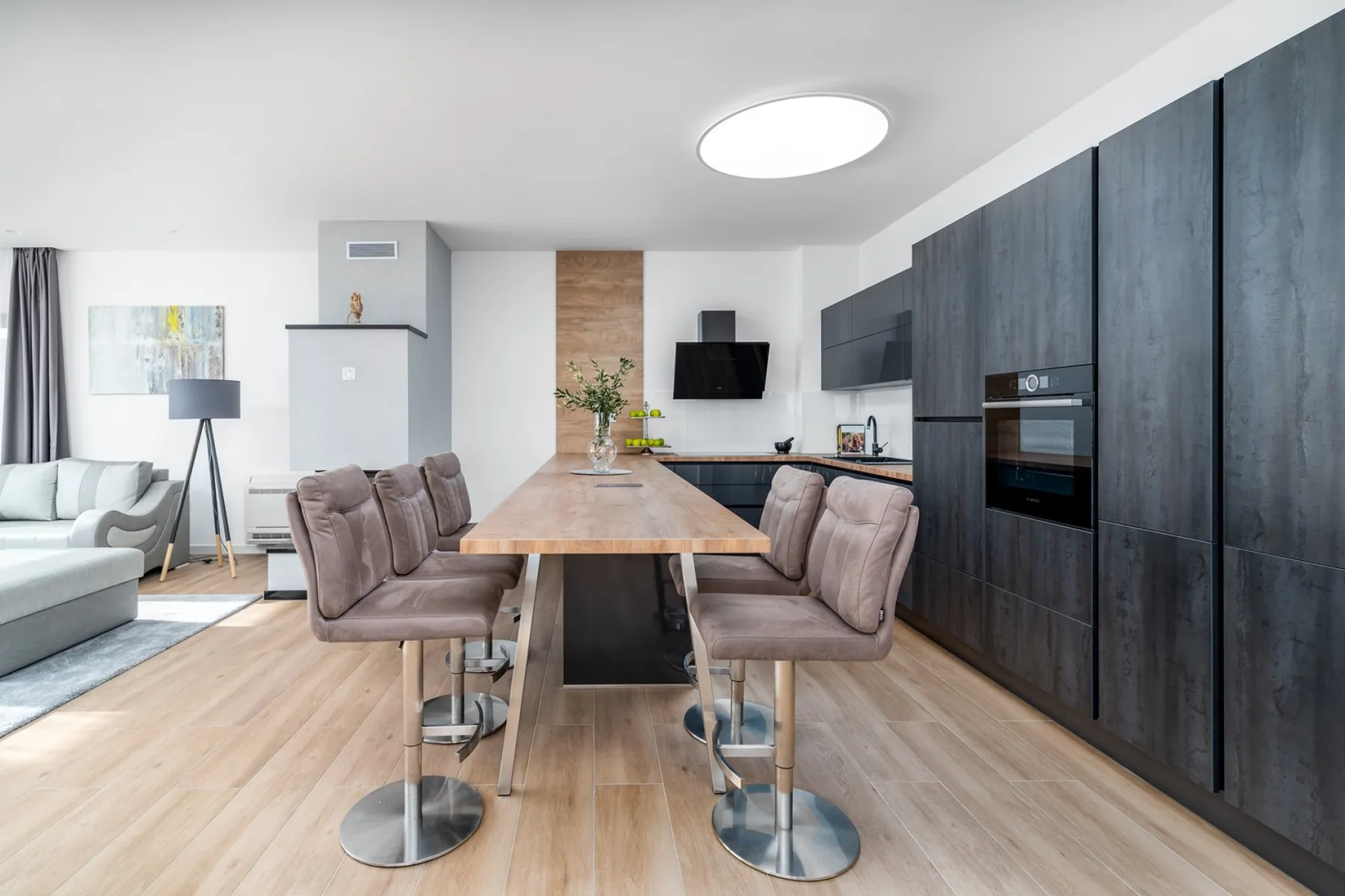 An open-plan dining area has a long wooden table, upholstered chairs, and a dark modern kitchen.