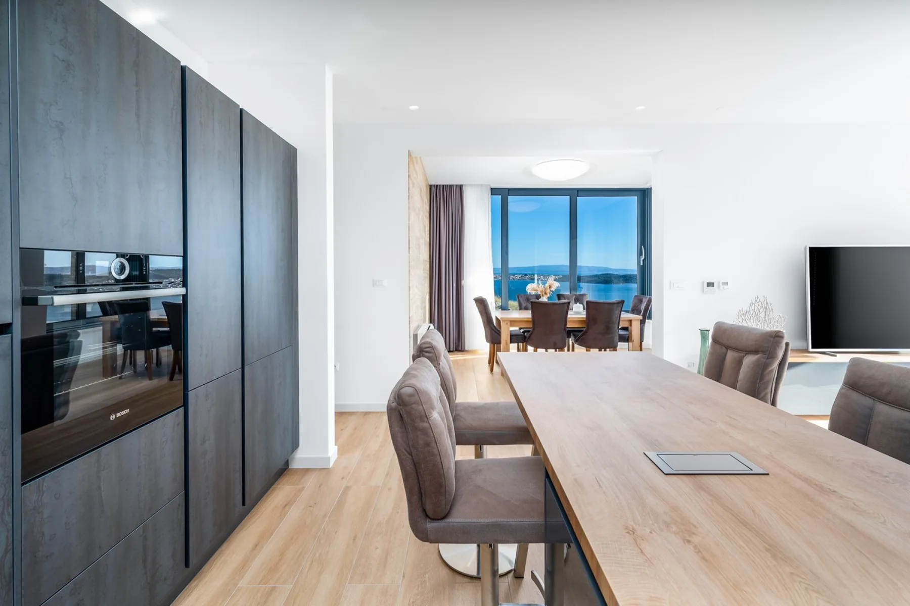 A modern dining area features a long wooden table, upholstered chairs, dark cabinetry, and large windows overlooking the sea.