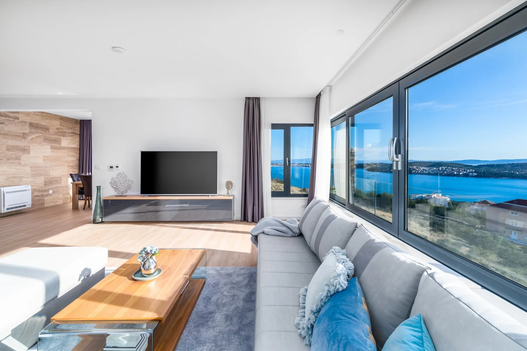 Bright living room with a gray sectional sofa, television, and broad windows overlooking the sea.