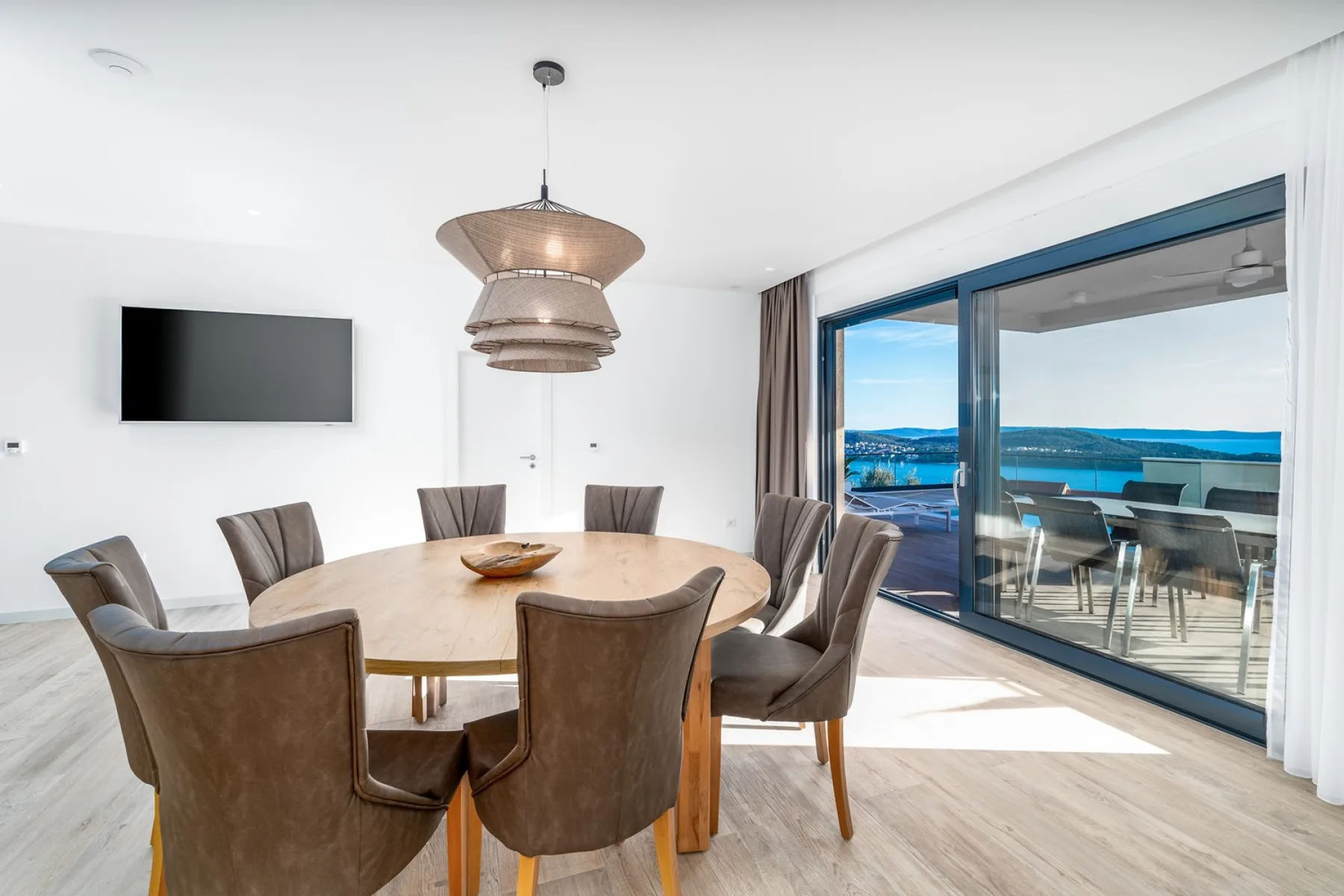 A dining area has a round table, upholstered chairs, a wall-mounted TV, and sliding glass doors overlooking the water.