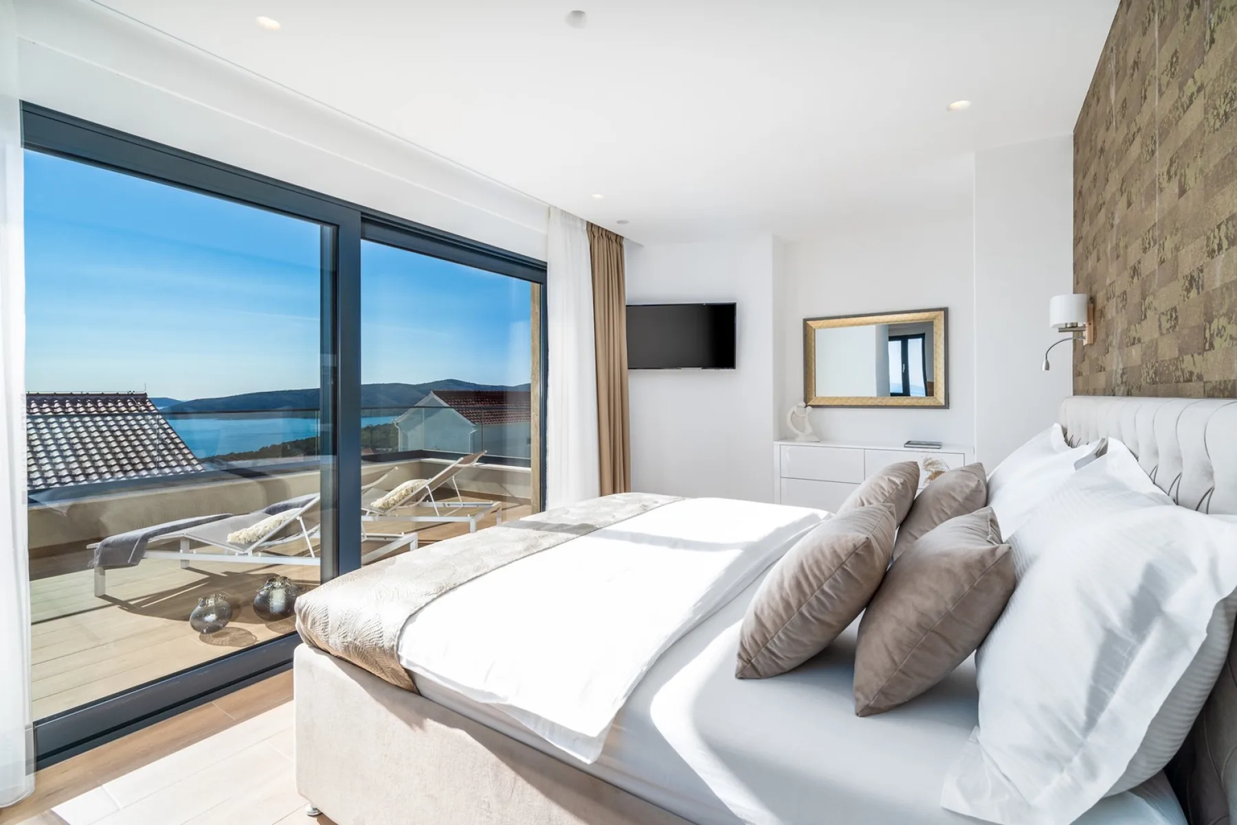 A bright bedroom features a large bed, floor-to-ceiling glass doors, and a view of the sea.