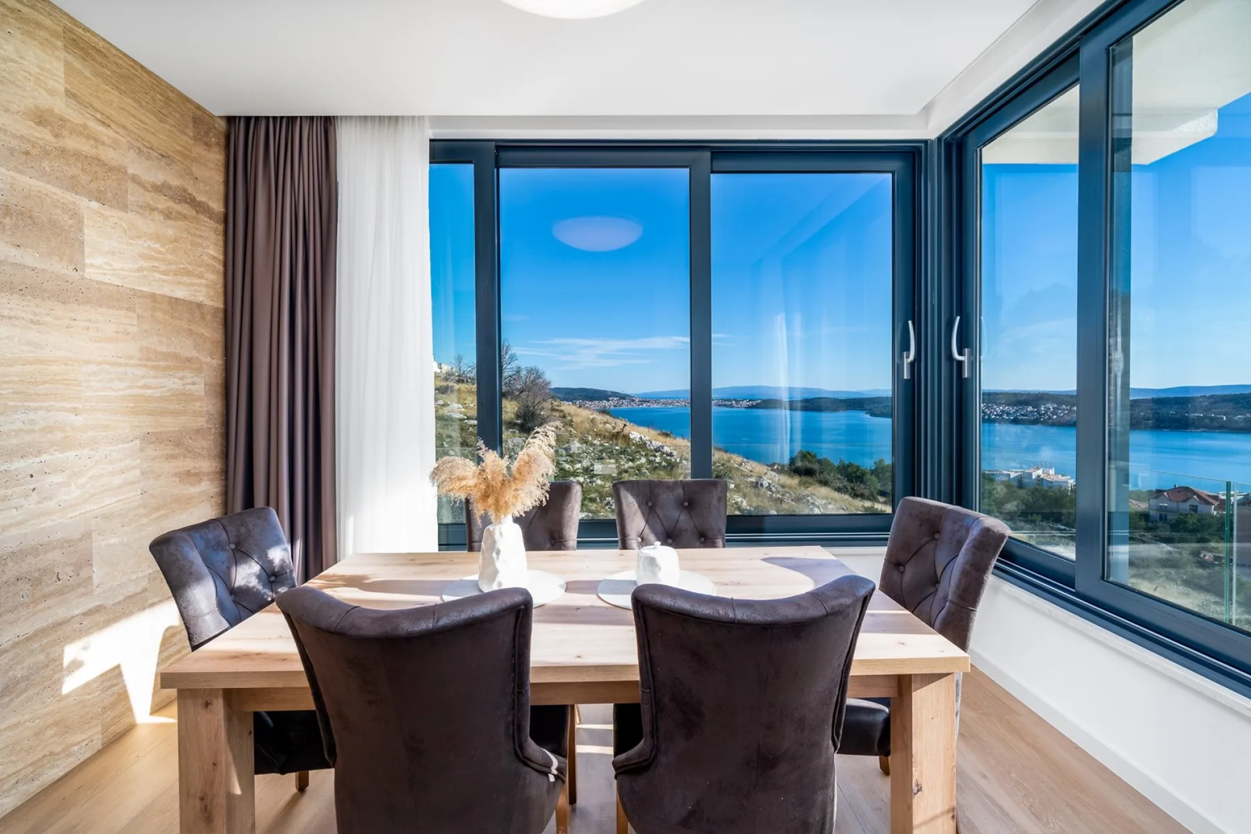 A dining area with a wooden table and chairs beside large windows overlooking the sea.