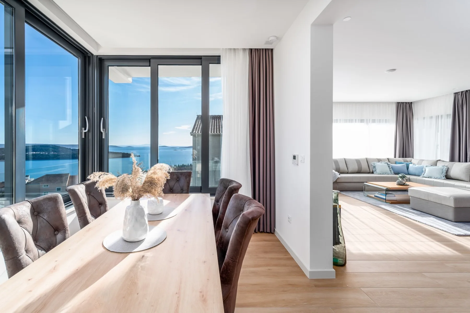 A bright dining area with a long wooden table and chairs beside large windows overlooking the sea.