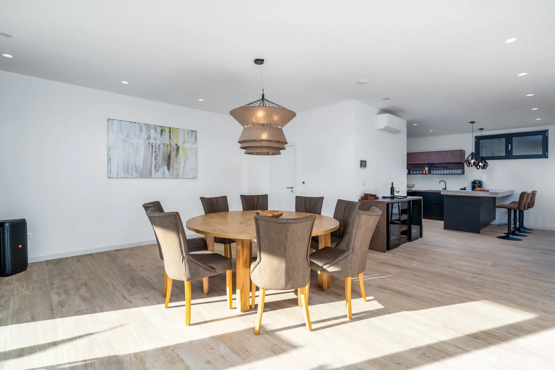 An open-plan dining and kitchen area with a round wooden table, upholstered chairs, and a dark kitchen island.