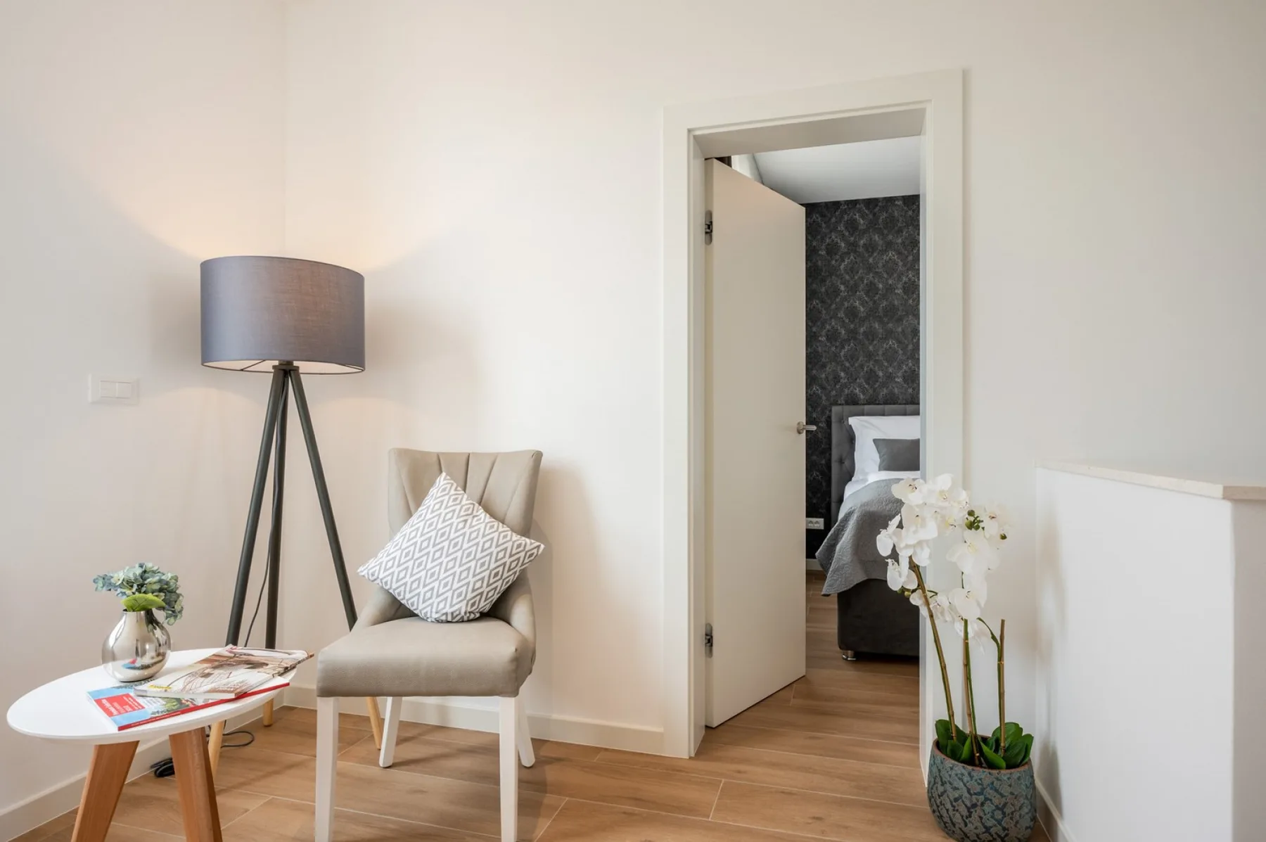 A bright sitting area has a beige chair, a tripod floor lamp, a small table, and an open doorway to a bedroom.