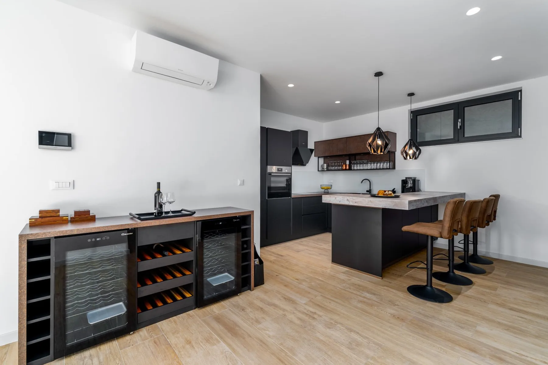 A modern kitchen features a central island with bar stools, dark cabinetry, pendant lights, and a wine cooler.