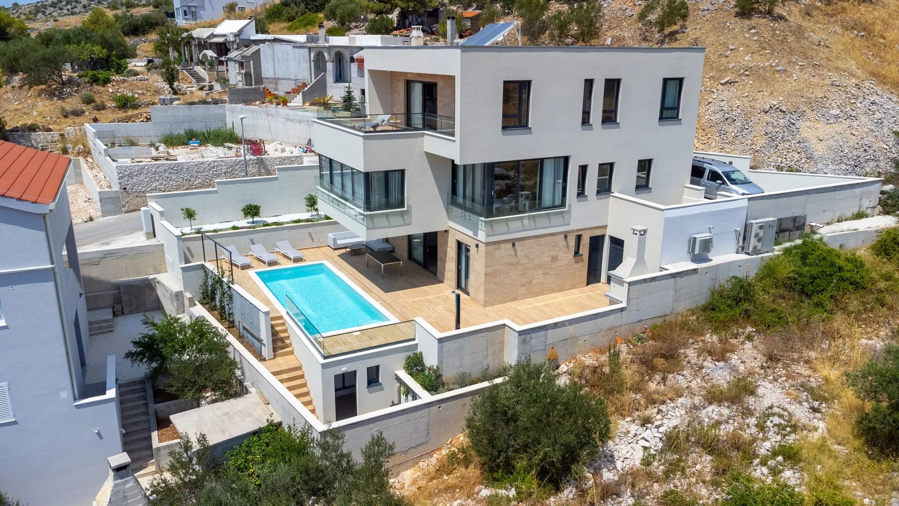 A modern white multi-level house overlooks a rectangular swimming pool and paved patio.