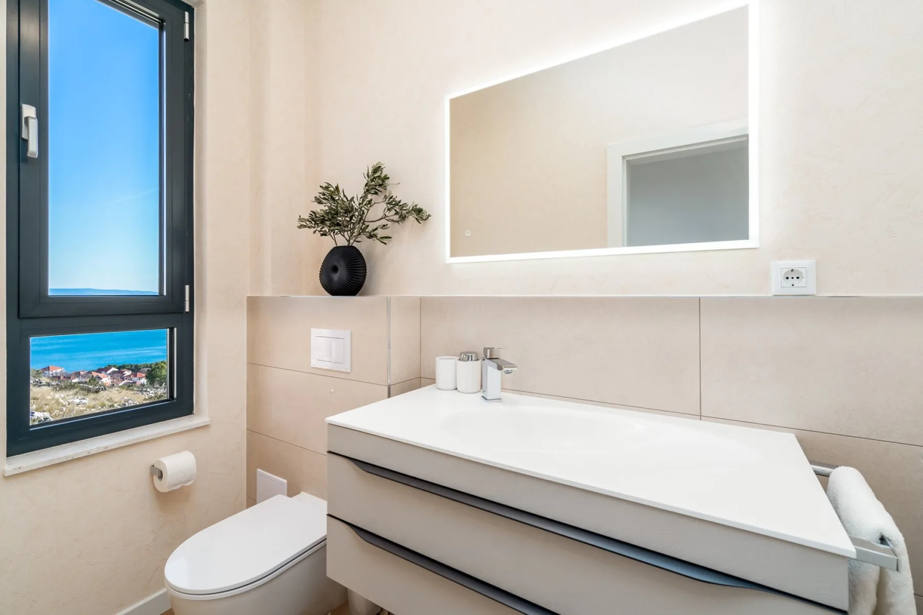 A bright bathroom has a wall-mounted vanity, rectangular mirror, toilet, and a window overlooking the sea.