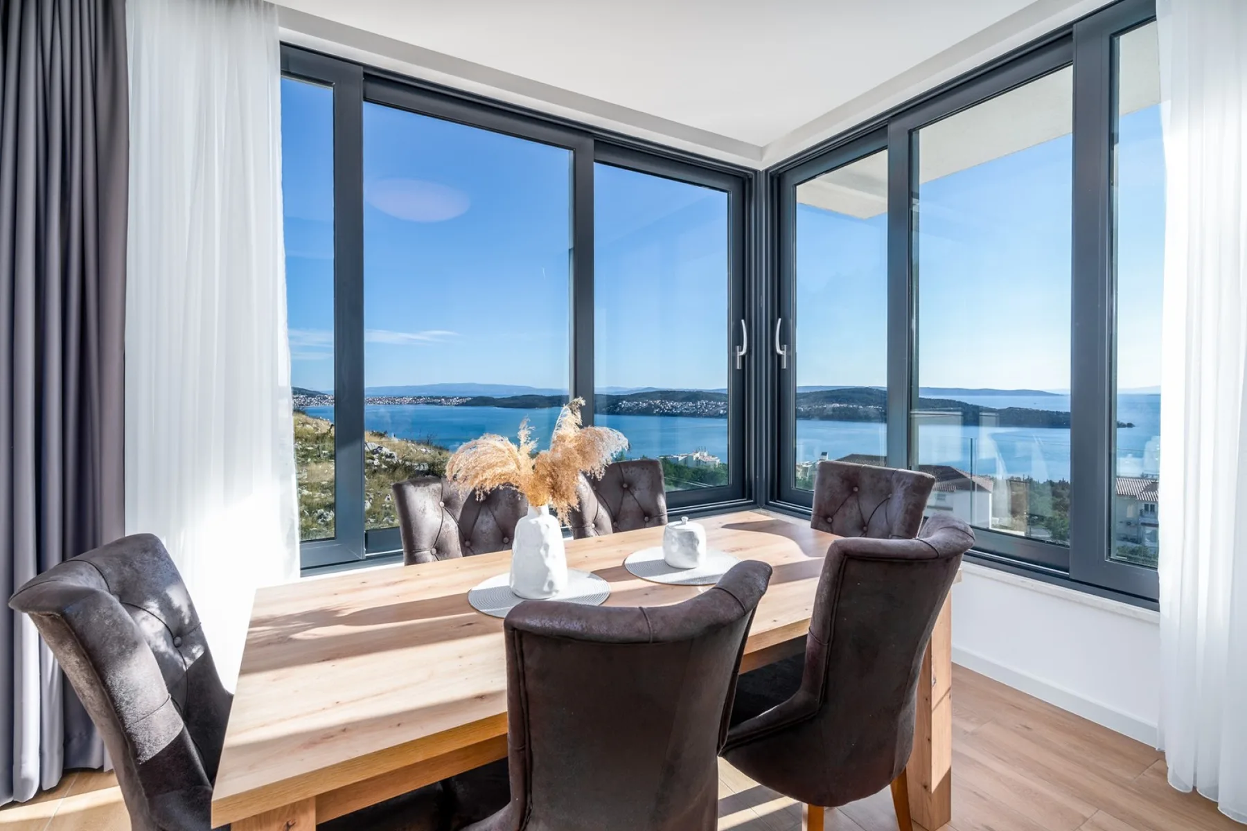 A wooden dining table and upholstered chairs sit beside floor-to-ceiling windows overlooking the water.
