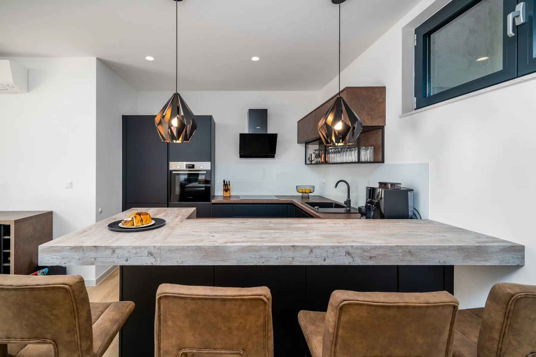 A modern kitchen has a broad stone island, dark cabinetry, pendant lights, and brown bar stools.