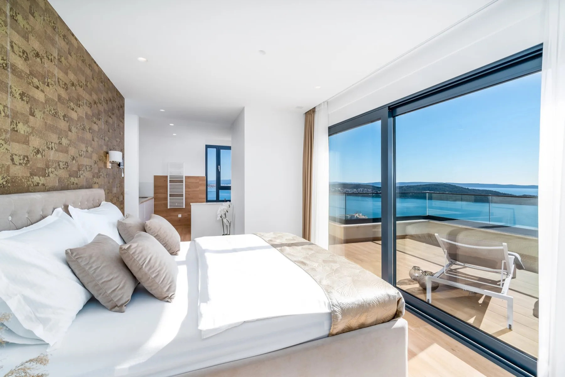 A bright bedroom has a large bed beside floor-to-ceiling glass doors opening onto a sea-view terrace.