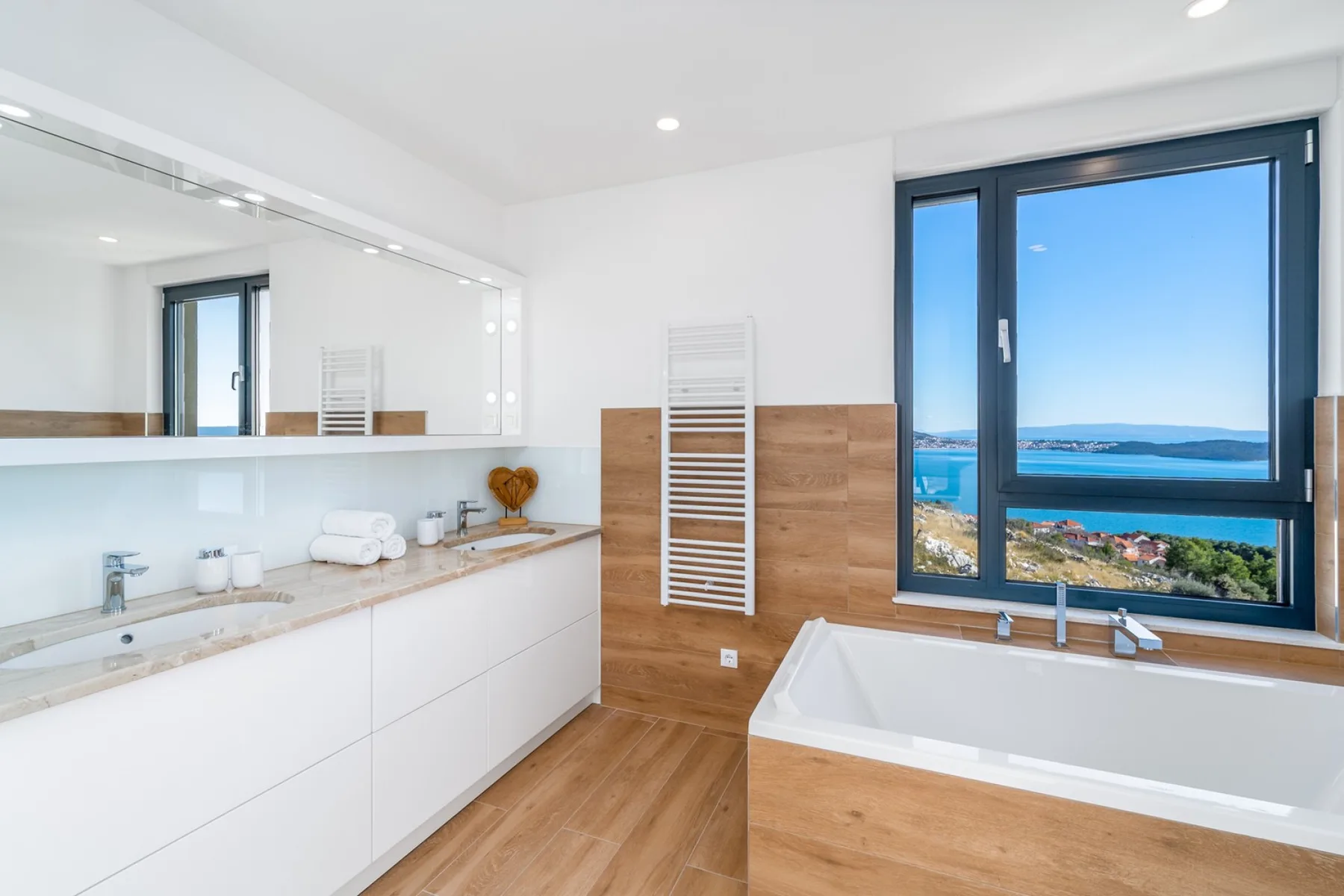 A bright bathroom has a wide vanity, a soaking tub, wood accents, and a large window overlooking the sea.