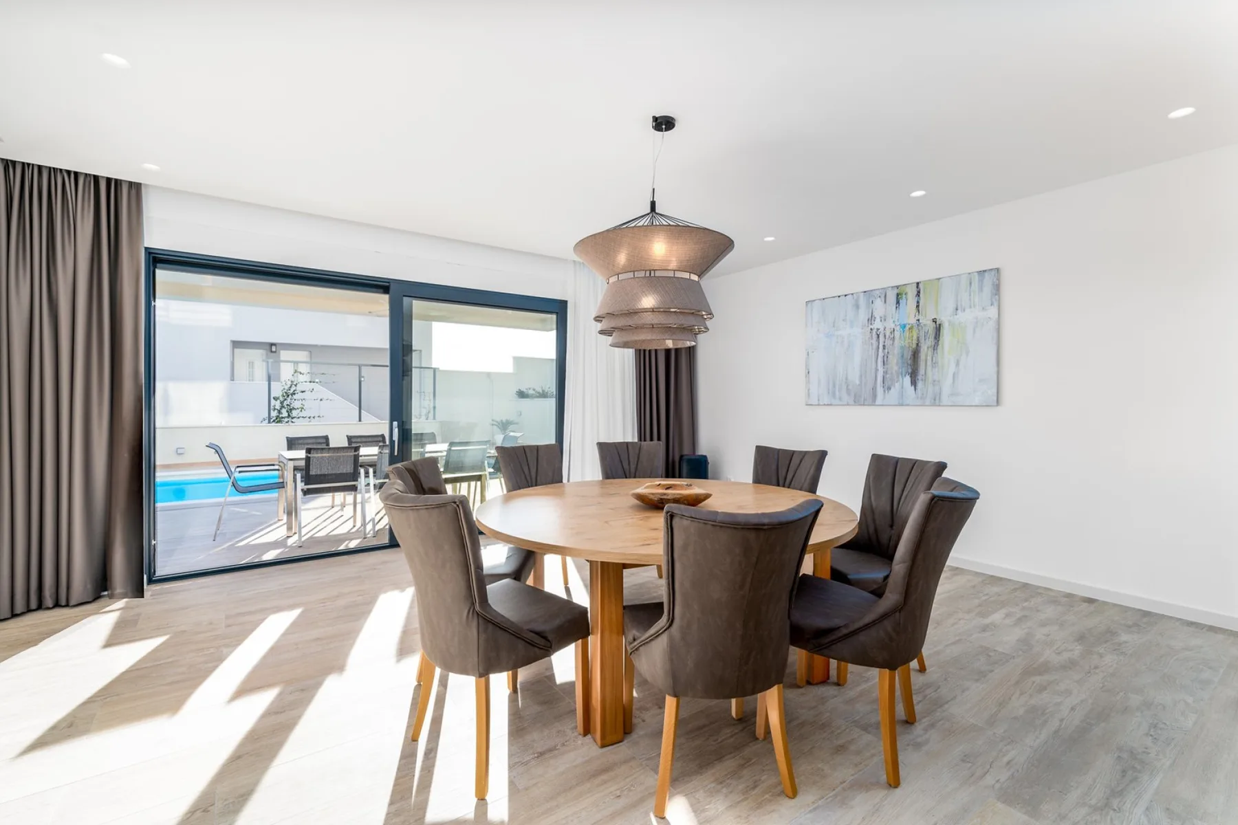 A dining room with a round wooden table, upholstered chairs, a pendant light, and sliding glass doors.
