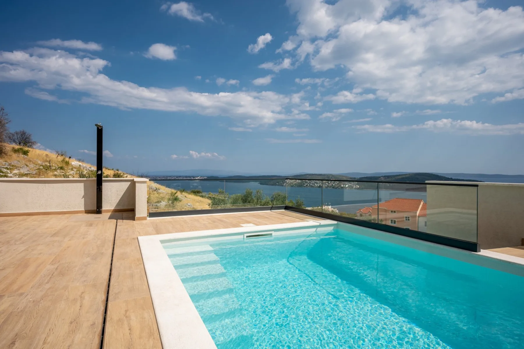 A rooftop pool sits beside a broad sunlit deck, with glass railings overlooking the water.