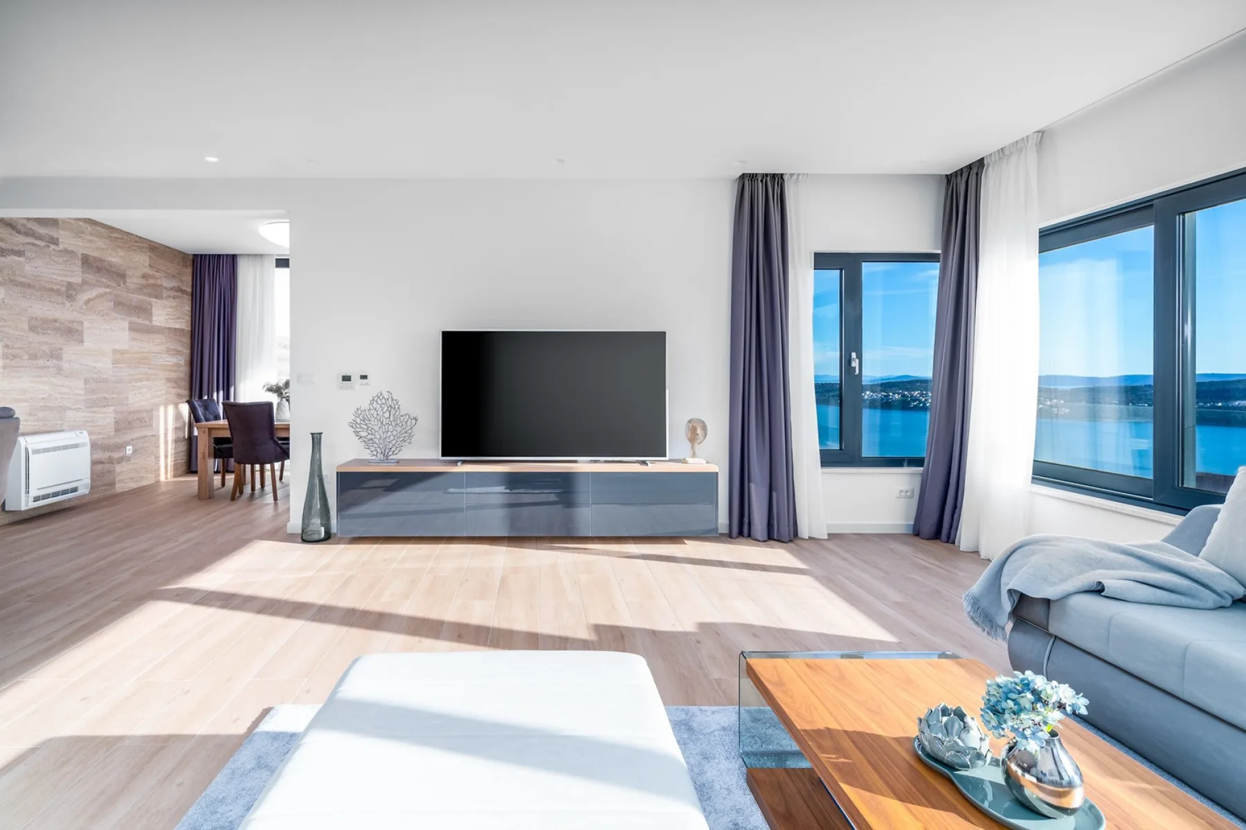 A bright living room features a television, blue sofa, wood coffee table, and large windows overlooking the water.