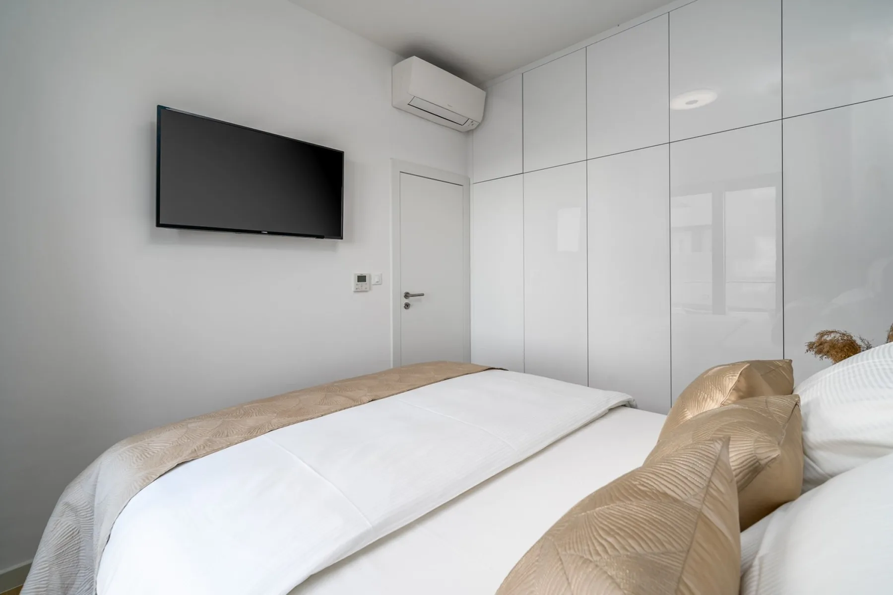 A bright bedroom with a white bed, beige pillows, a wall-mounted television, and built-in wardrobes.