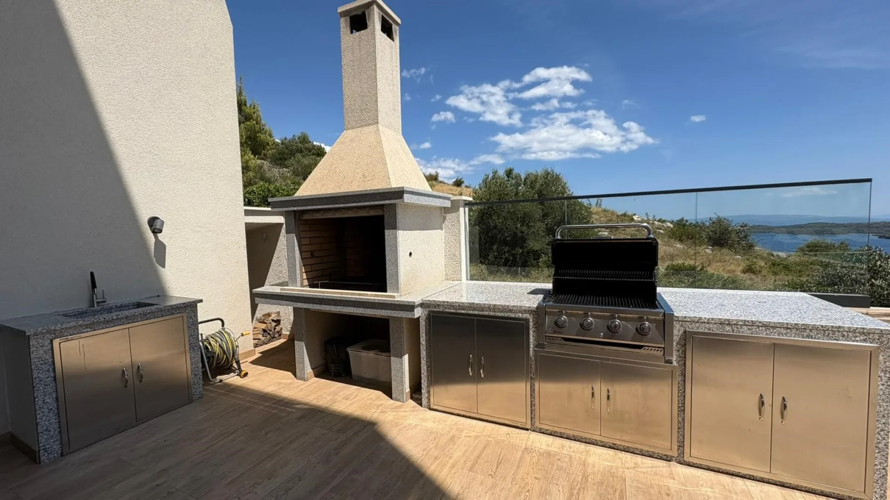 An outdoor kitchen features a stone barbecue, stainless-steel cabinets, and a glass railing overlooking the water.