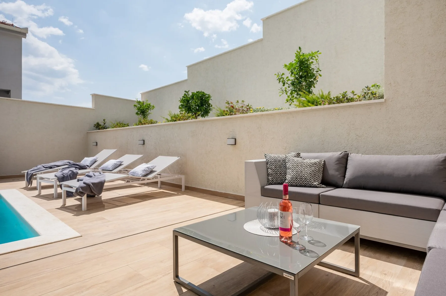 A sunny rooftop terrace has a gray outdoor sofa, glass coffee table, lounge chairs, and a small pool.