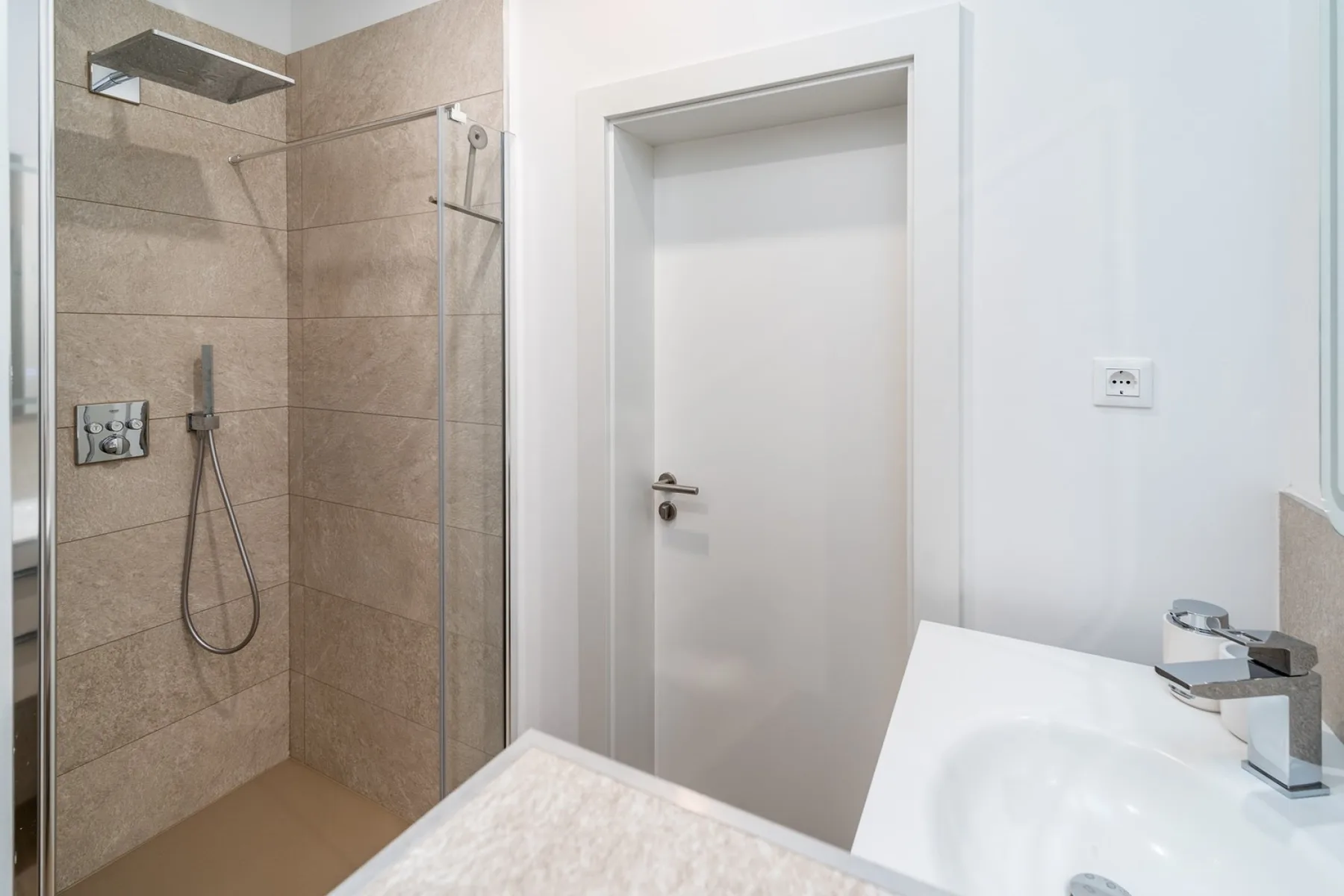 A modern bathroom with a glass shower, beige tile, a white door, and a sink.
