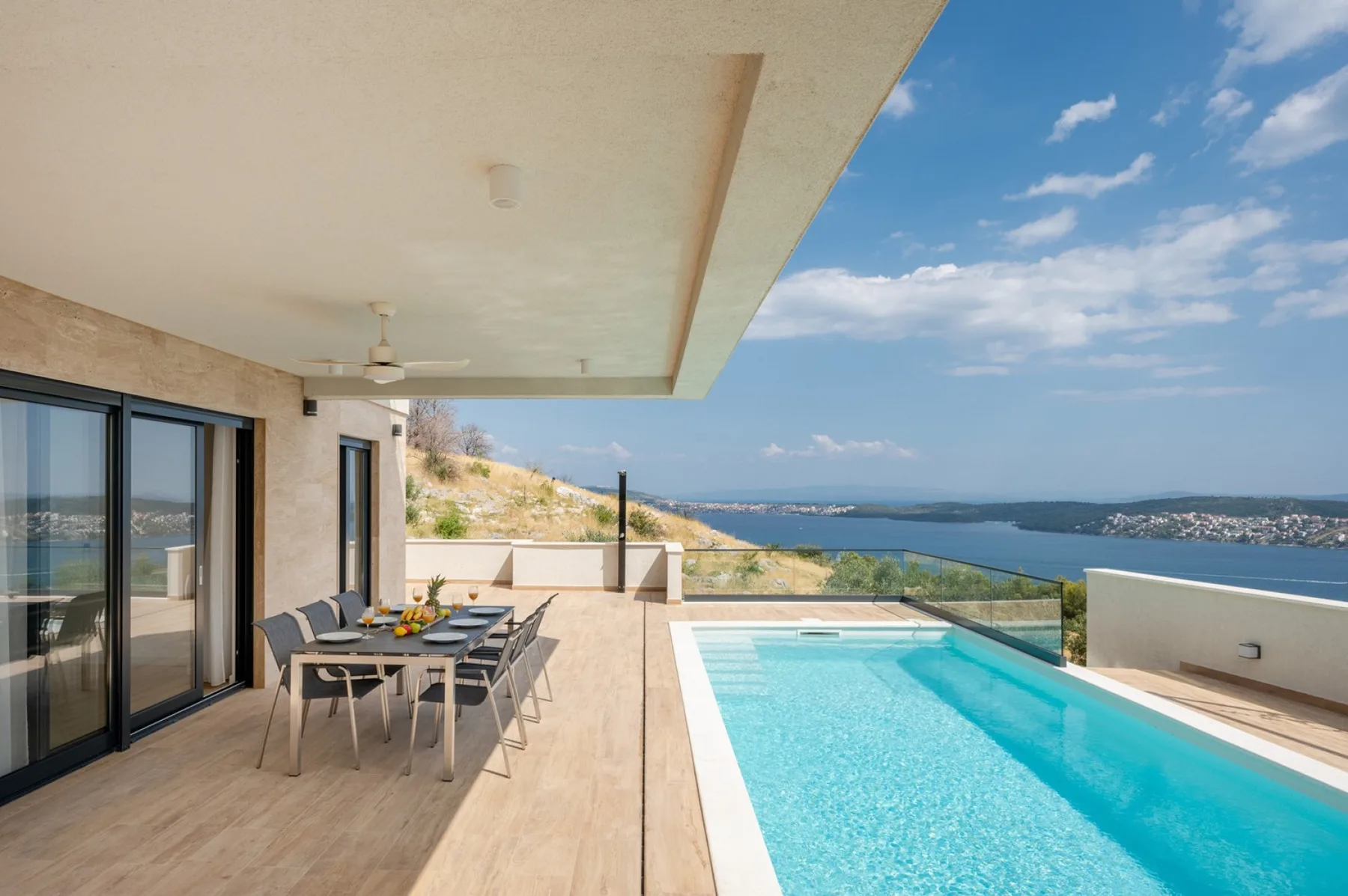 A sunny terrace features a long swimming pool, outdoor dining table, and expansive coastal views.