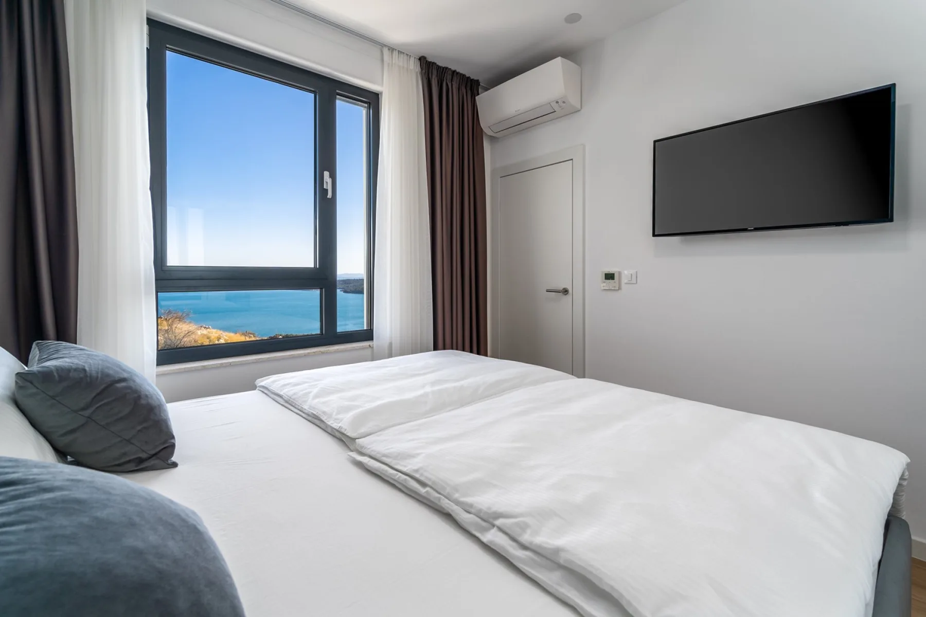 A bright bedroom with a neatly made bed, wall-mounted television, and ocean views through the window.