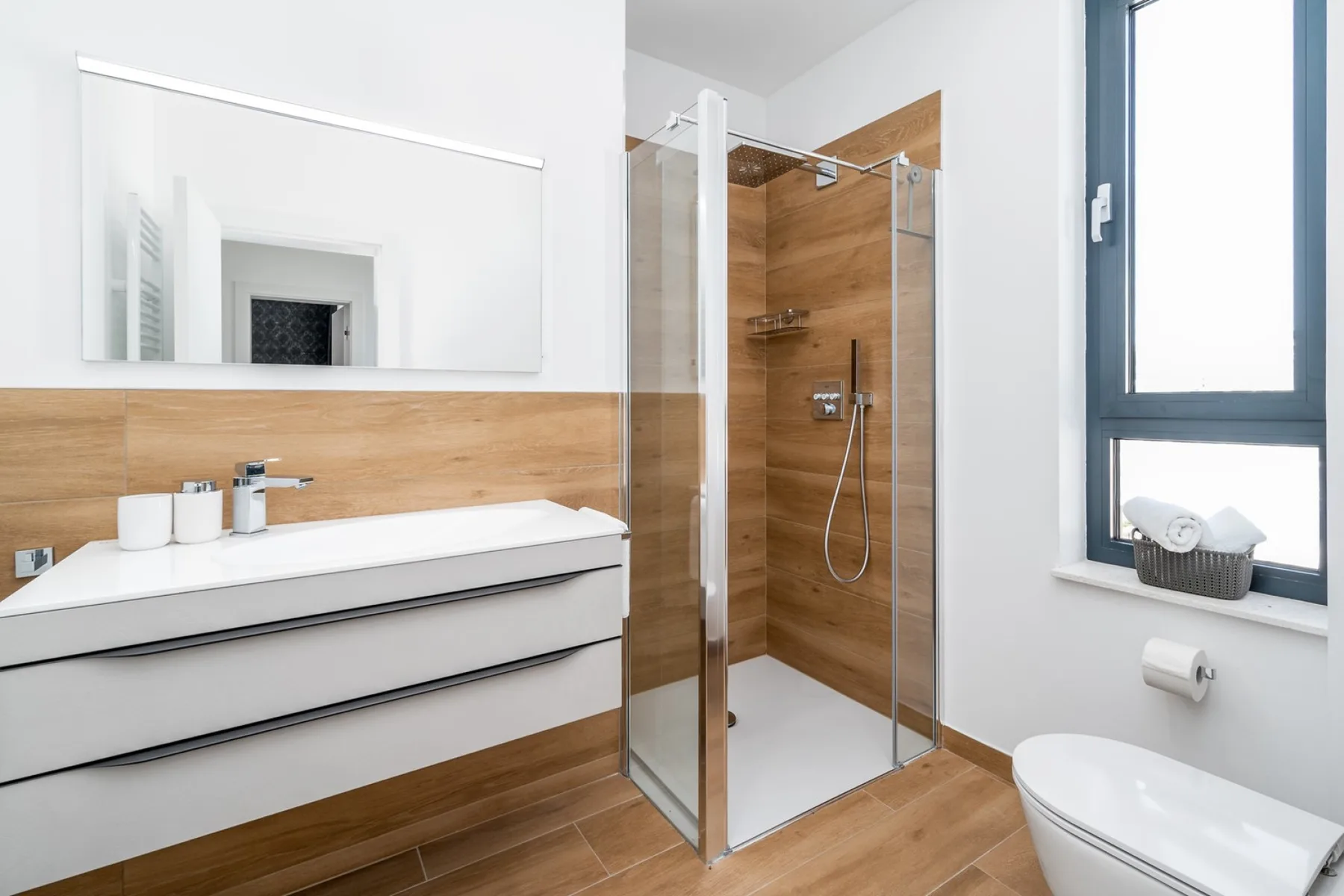 A modern bathroom features a wide vanity, a glass-enclosed shower with wood-look tile, and a large window.