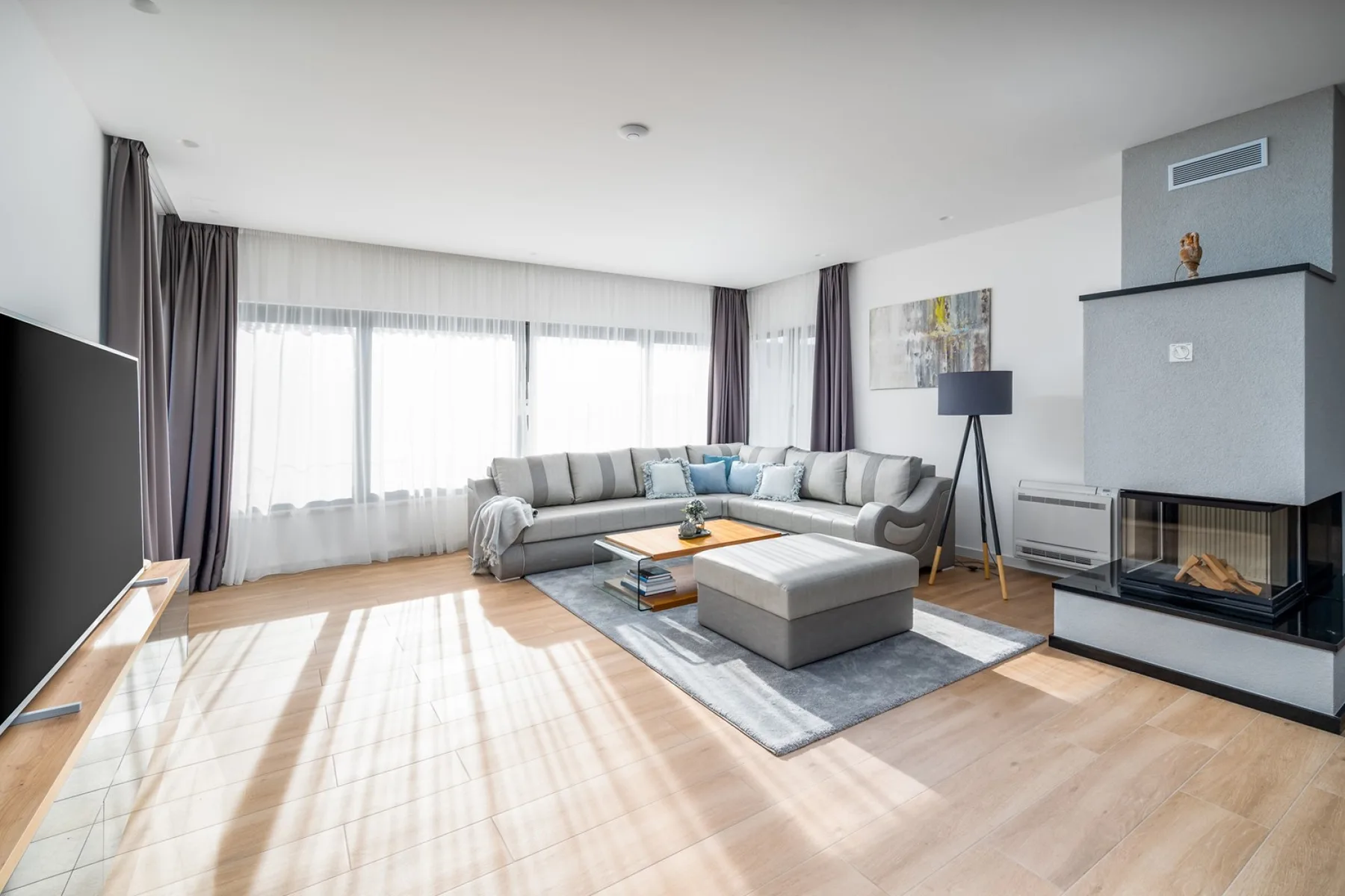 A bright living room has an L-shaped gray sofa, ottoman, large windows, and a modern fireplace.