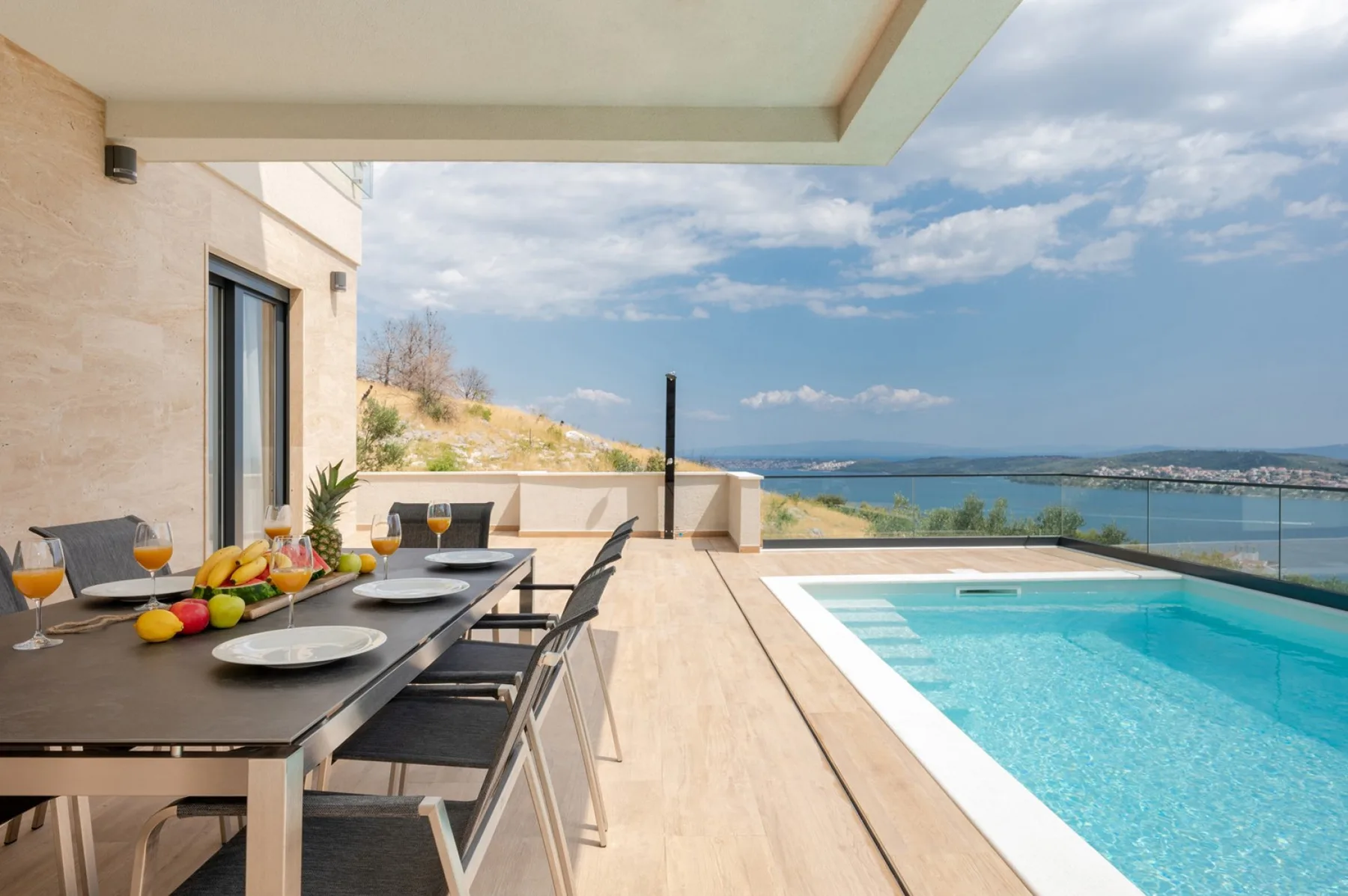 A covered terrace has a dining table and chairs beside a swimming pool overlooking the coast.