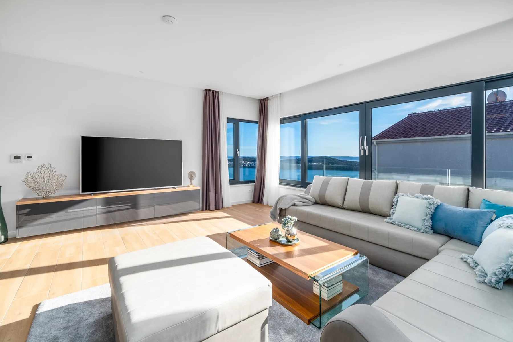 A bright living room features a gray sectional sofa, a large television, and floor-to-ceiling windows overlooking the water.