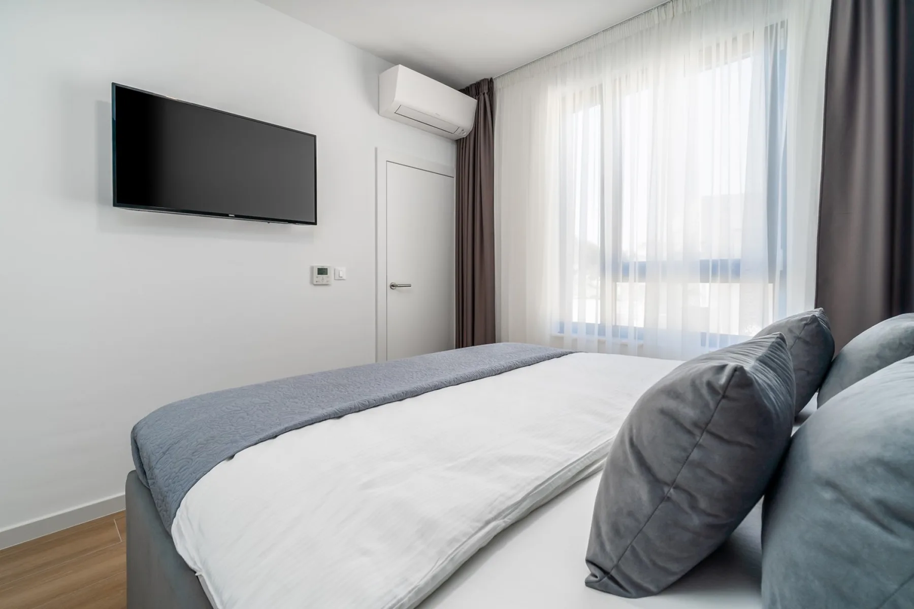 A bedroom with a neatly made bed, wall-mounted television, and a large curtained window.