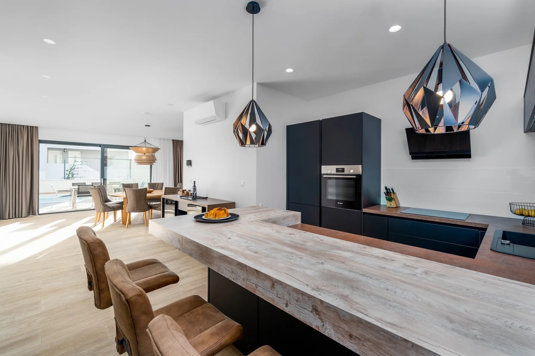 A bright open-plan kitchen and dining area features a large stone island, dark cabinets, and pendant lights.